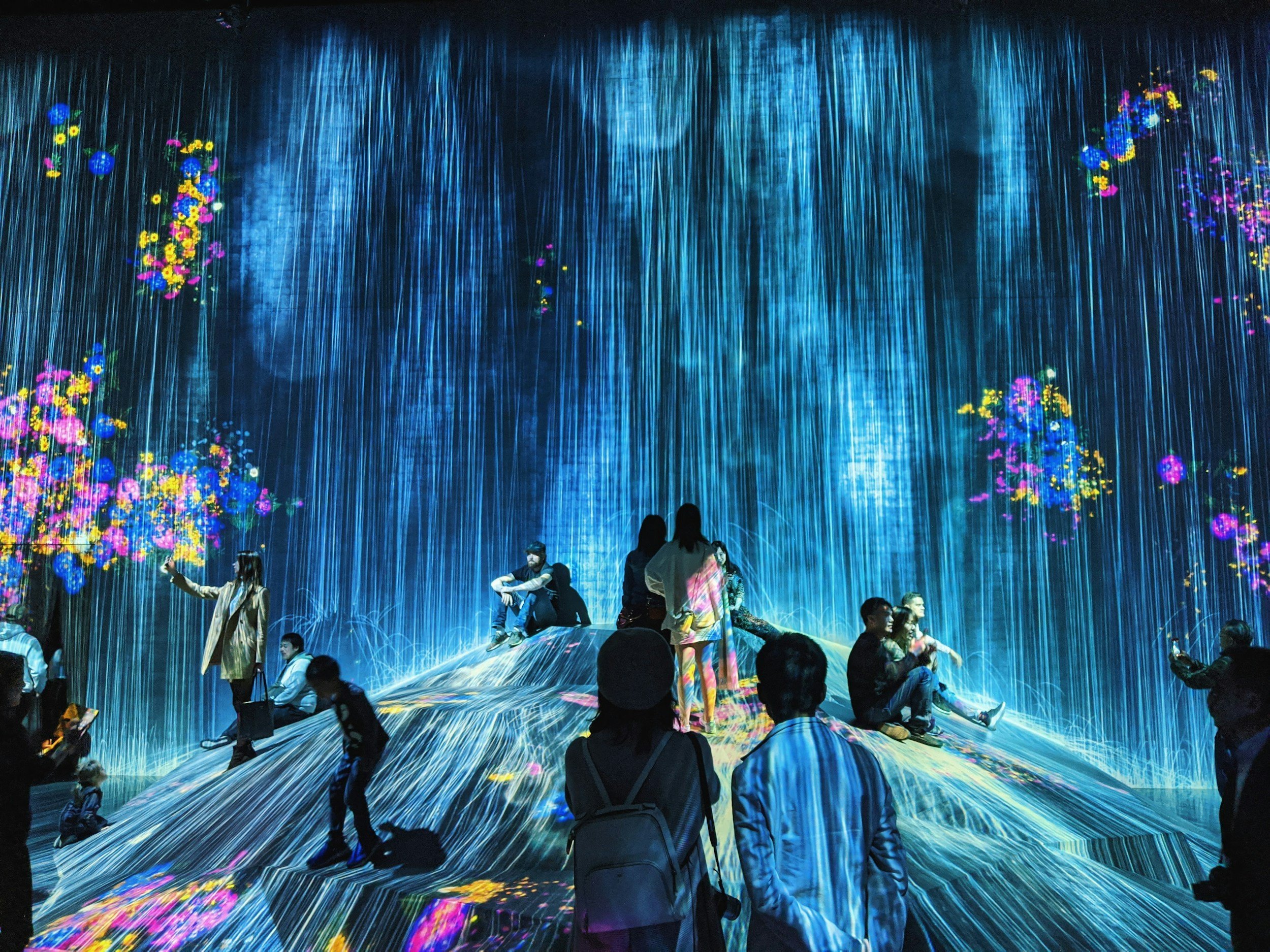 A vibrant digital art installation with a waterfall of blue light, surrounded by colorful flowers and multiple people observing and taking pictures.
