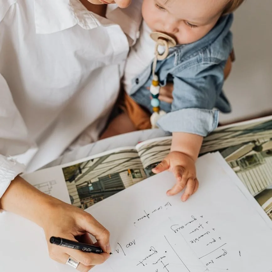 A woman and a young child looking at a large open photo album on a table, with the woman writing notes or a letter in a notebook.