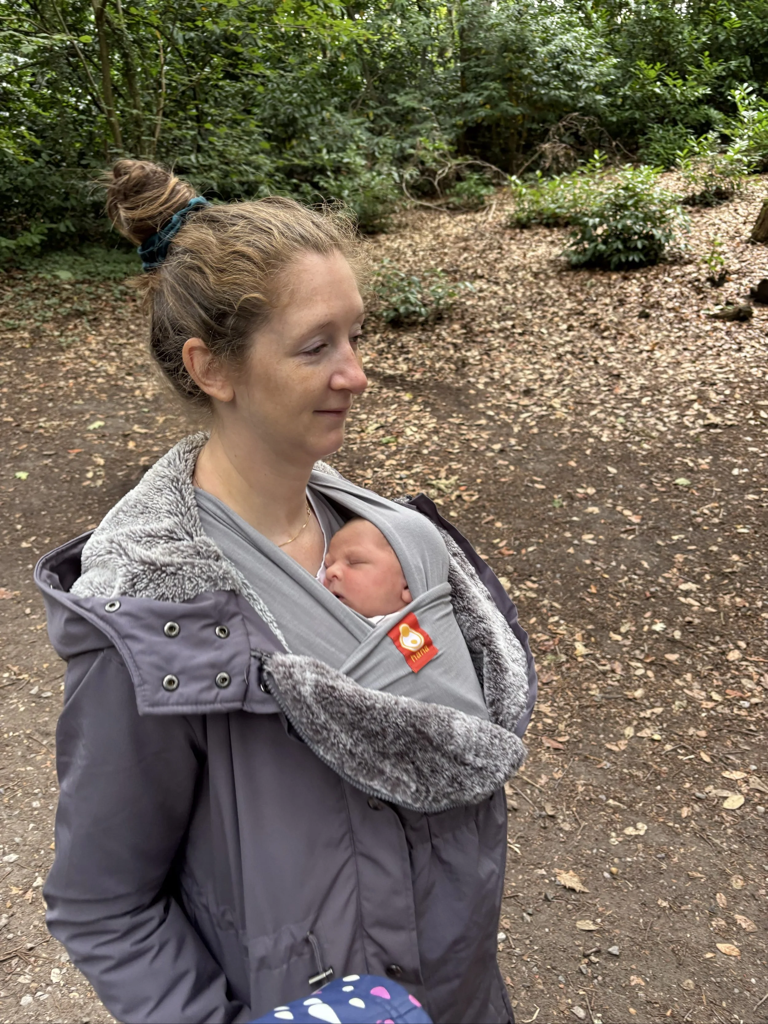 A woman with light brown hair tied in a bun, wearing a gray coat with a fur-lined hood, is walking outdoors in a wooded area while carrying a sleeping baby in a gray baby carrier.