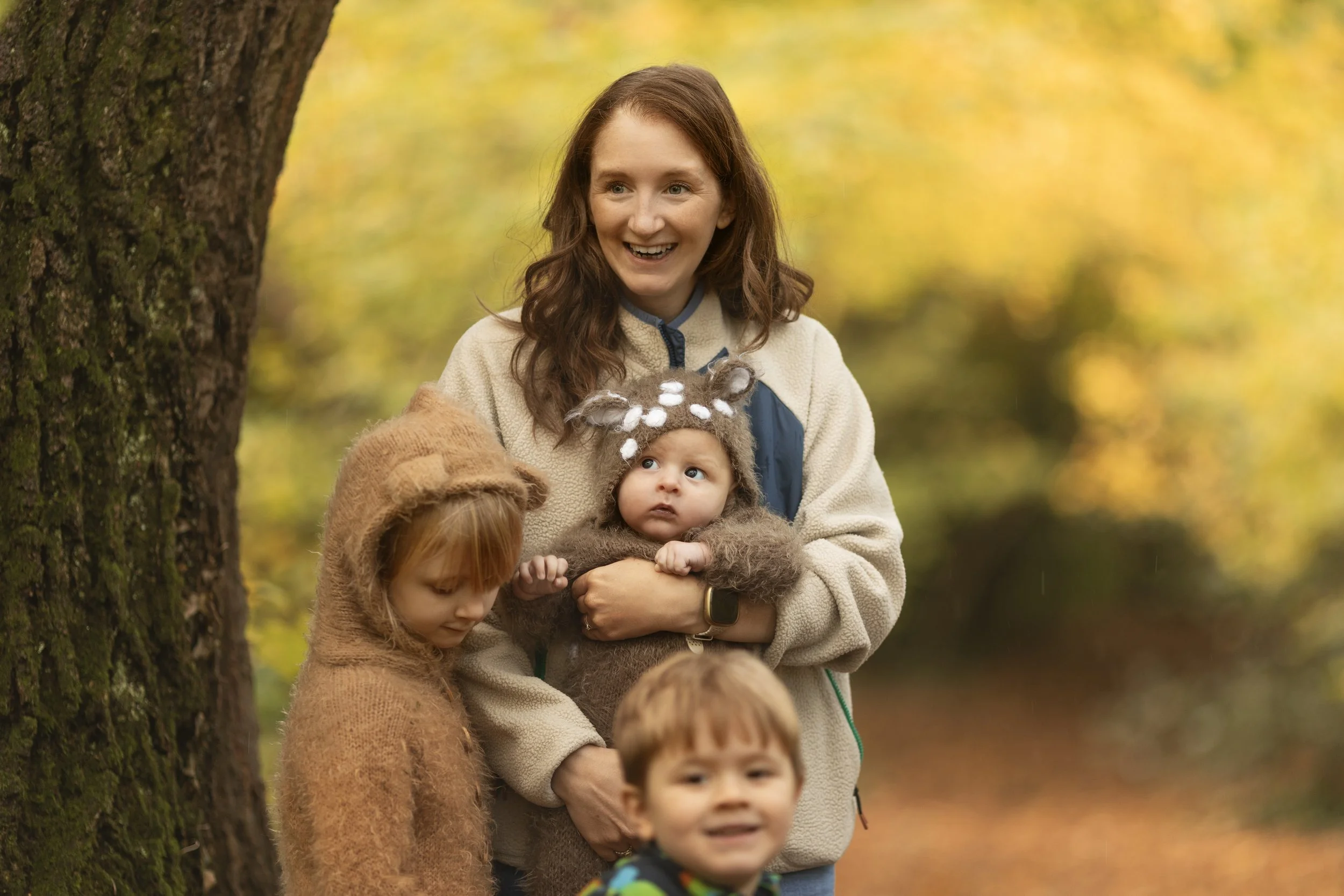 A woman with red hair smiling while holding a baby girl wearing a reindeer hat, standing among children in an autumn forest.