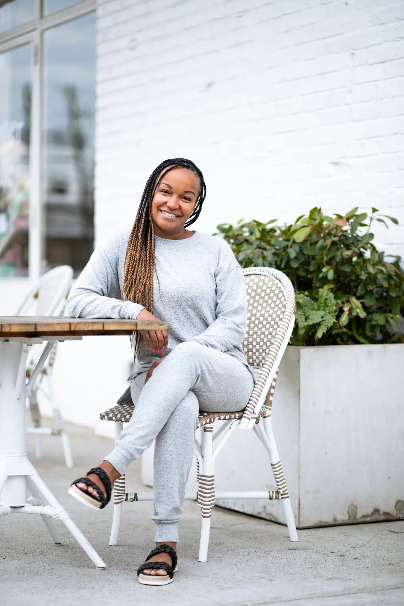 A woman with braided hair sitting outside at a table, smiling at the camera, wearing a gray sweatshirt and sweatpants.