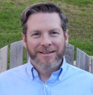 A man with a beard and short hair, wearing a light blue button-up shirt, smiling outdoors with grass and wooden fencing in the background.