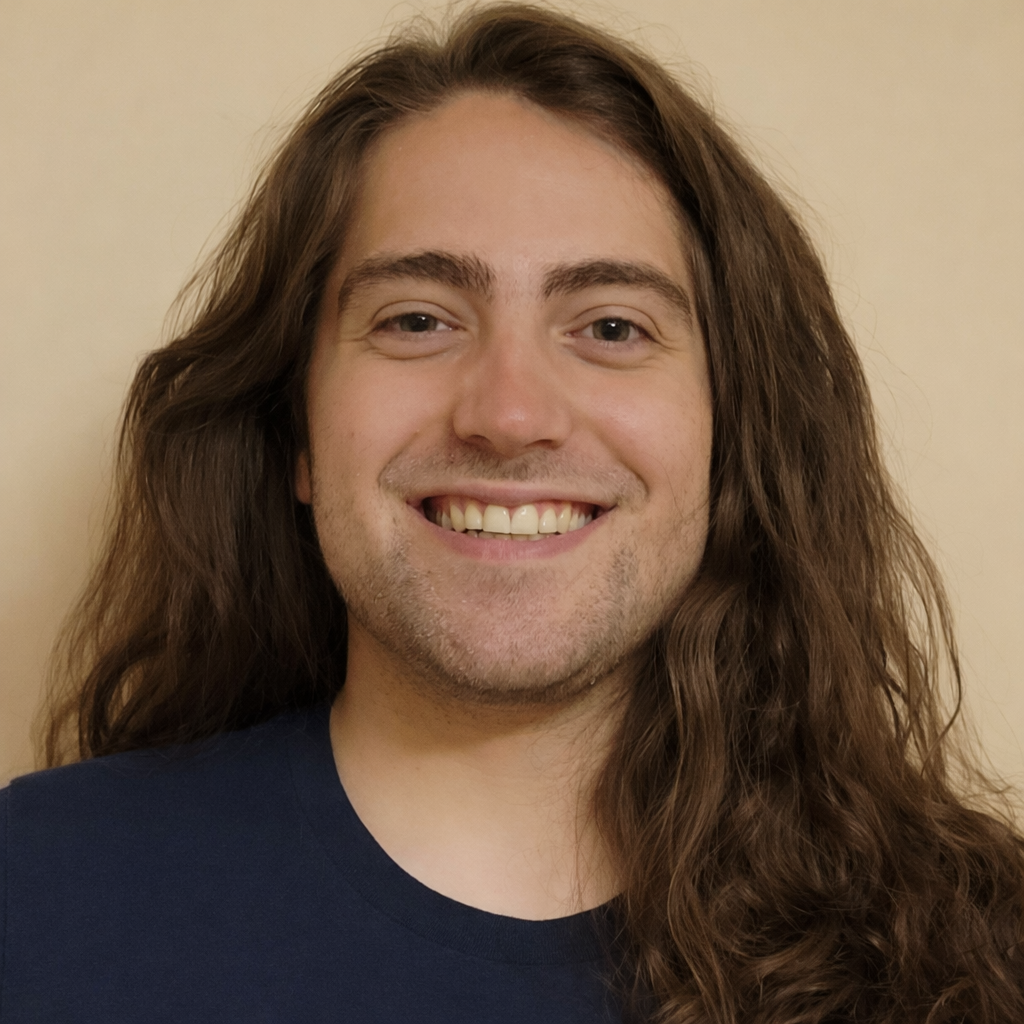 A smiling man with long brown hair, wearing a dark shirt, standing against a beige wall.