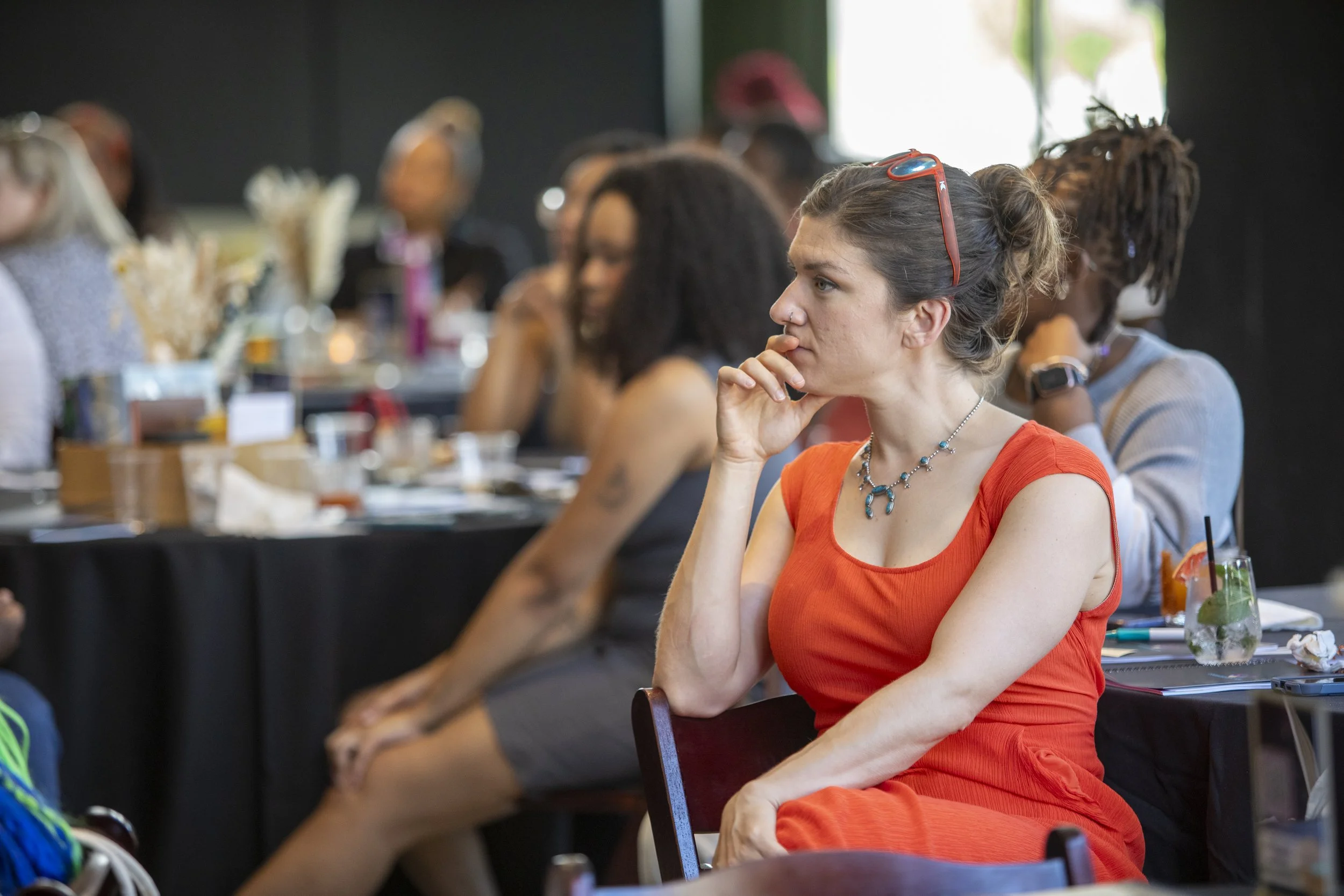 A woman with curly hair wearing a red dress and necklace, sitting at a table, appearing attentive at a conference or seminar. Multiple other attendees are seated behind her.