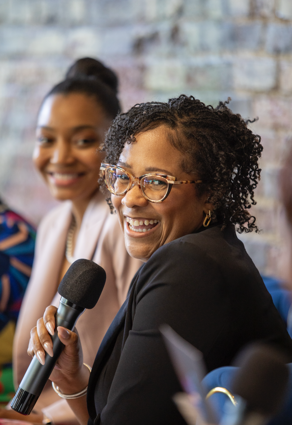 A woman with curly hair and glasses smiling while holding a microphone, sitting next to a woman with her hair styled in a bun, both in a discussion or panel setting.