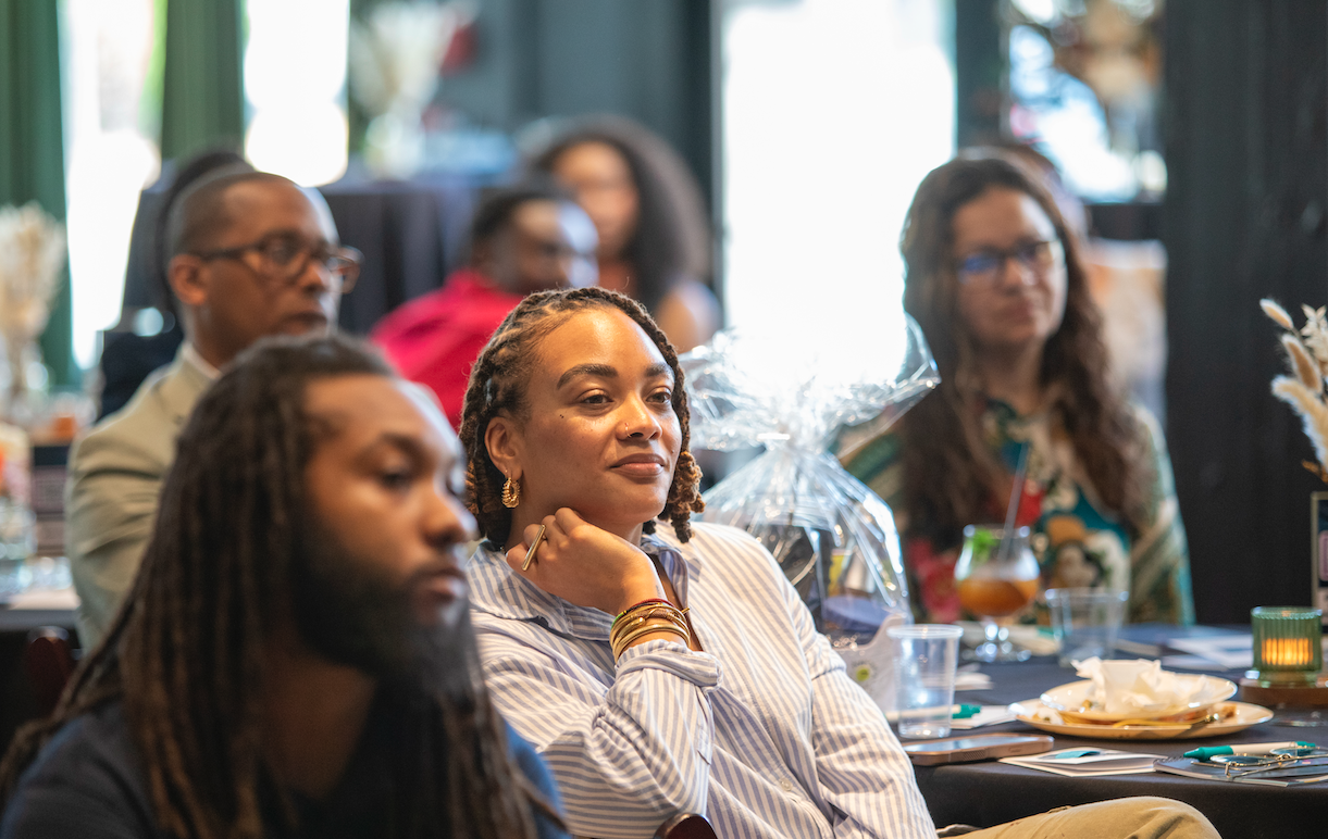 A group of people seated at a table during a gathering or event, with one woman in the center looking attentively, surrounded by others in a casual setting.