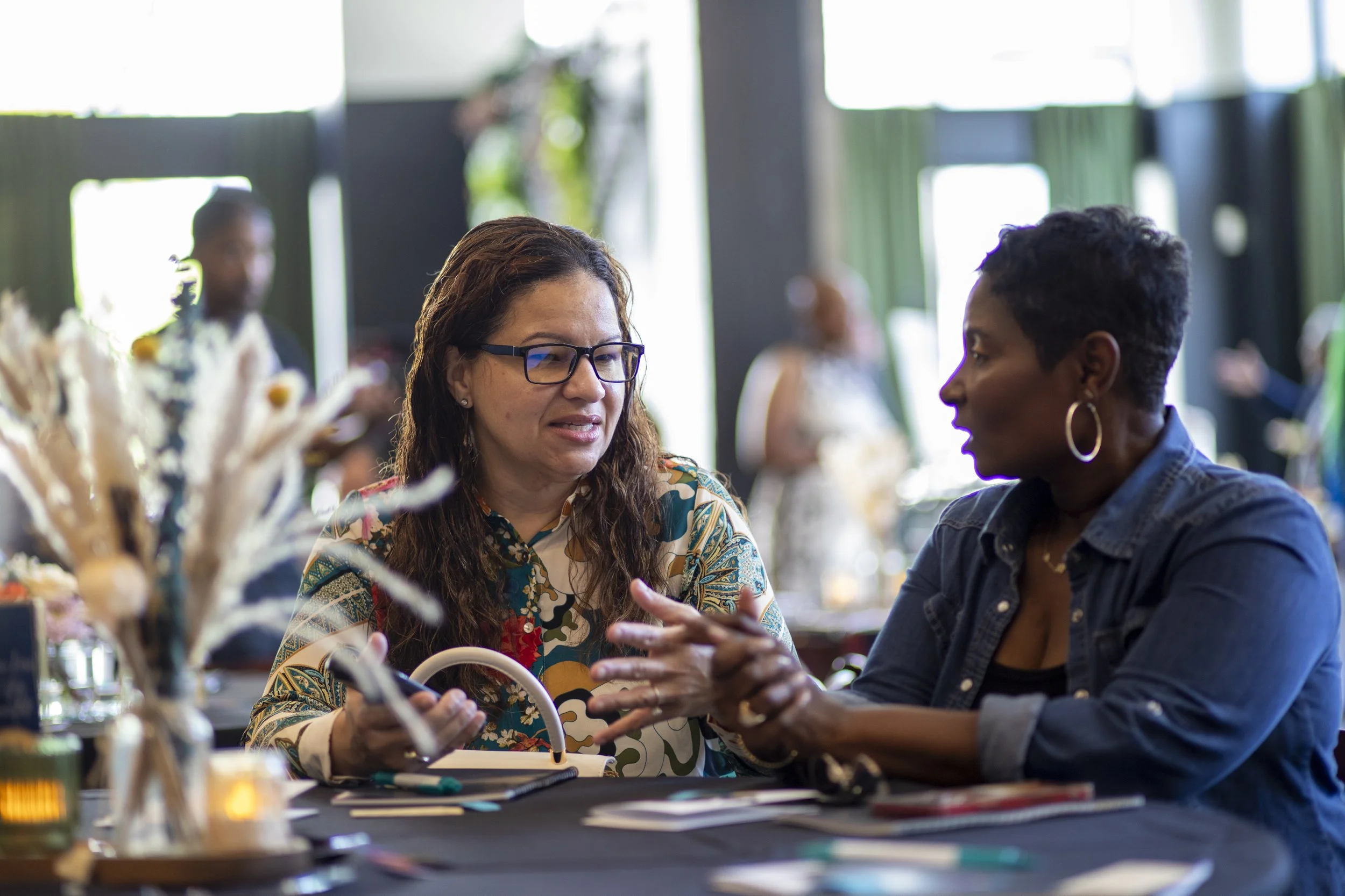 Two women sitting at a table engaged in conversation with others visible in the background at an indoor event.