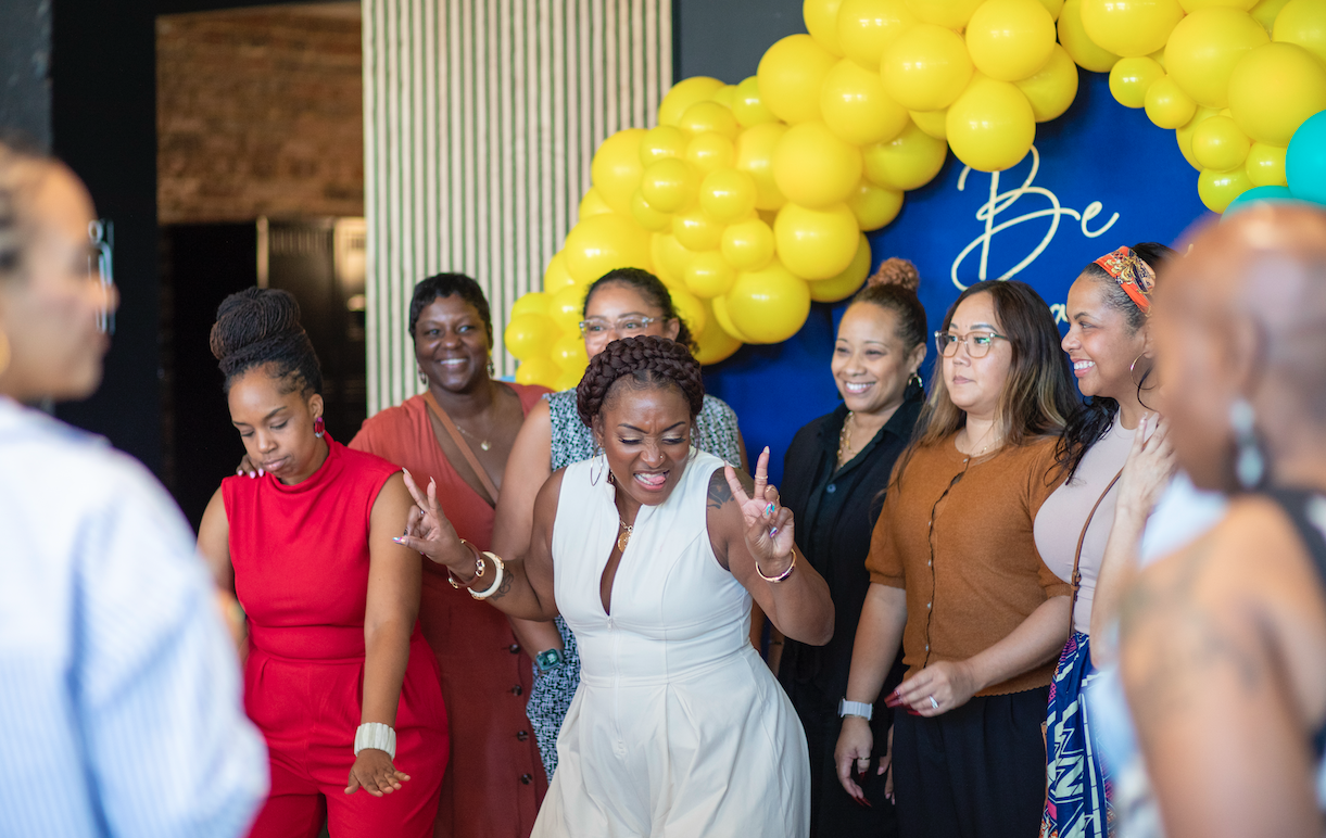 Group of women at a celebration or event, with yellow balloons in the background, some smiling and others engaged in conversation.