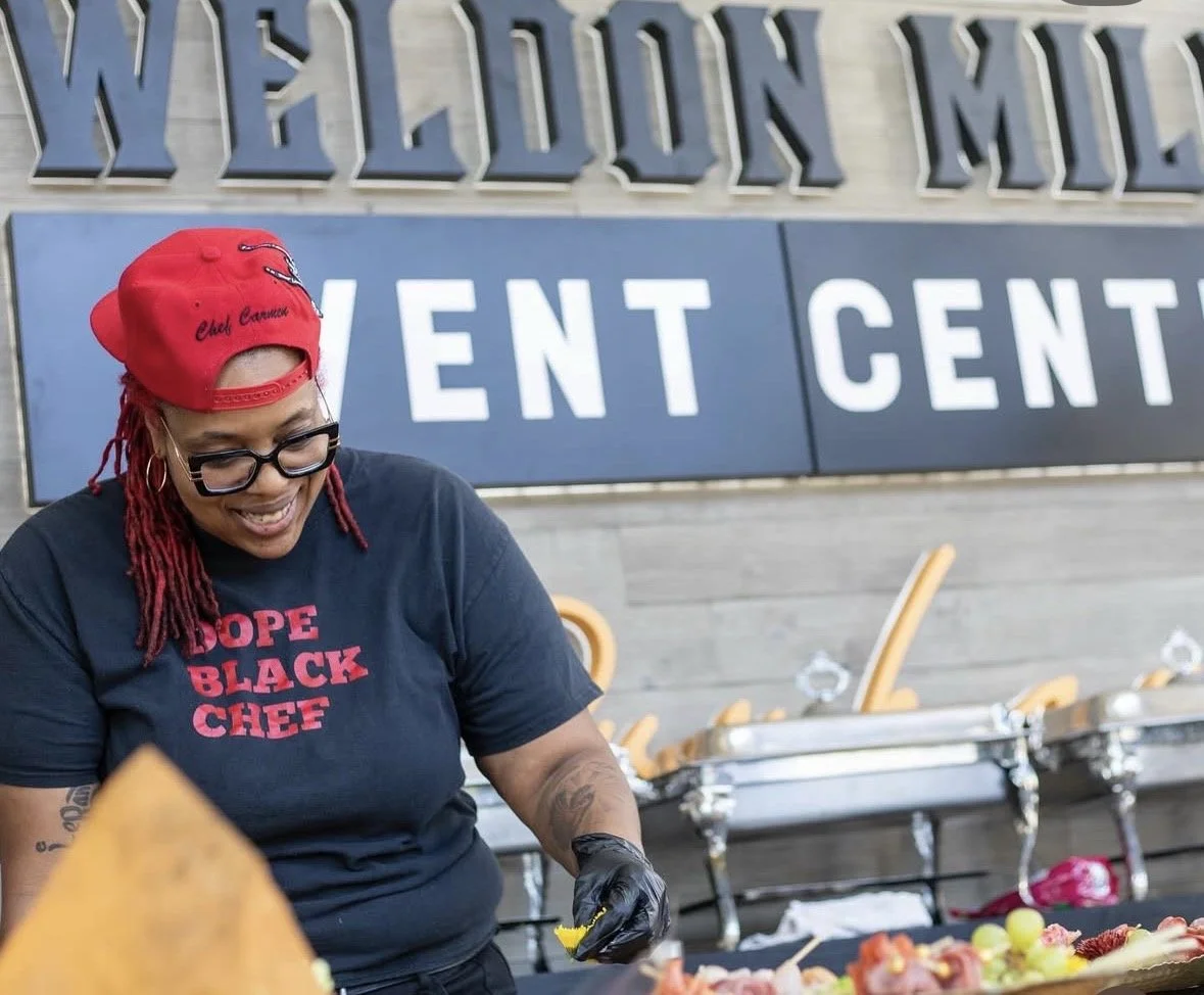 A woman with red dreadlocks wearing glasses, a red cap, and a black t-shirt with red text that reads 'Hope Black Chef' is smiling and preparing food at a food event. Behind her is a sign with large white letters spelling 'VENT' and part of a larger s