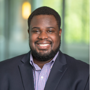 A smiling African American man with short hair and a beard, wearing a dark suit jacket and a light-colored checkered shirt, standing indoors with a blurred green and yellow background.