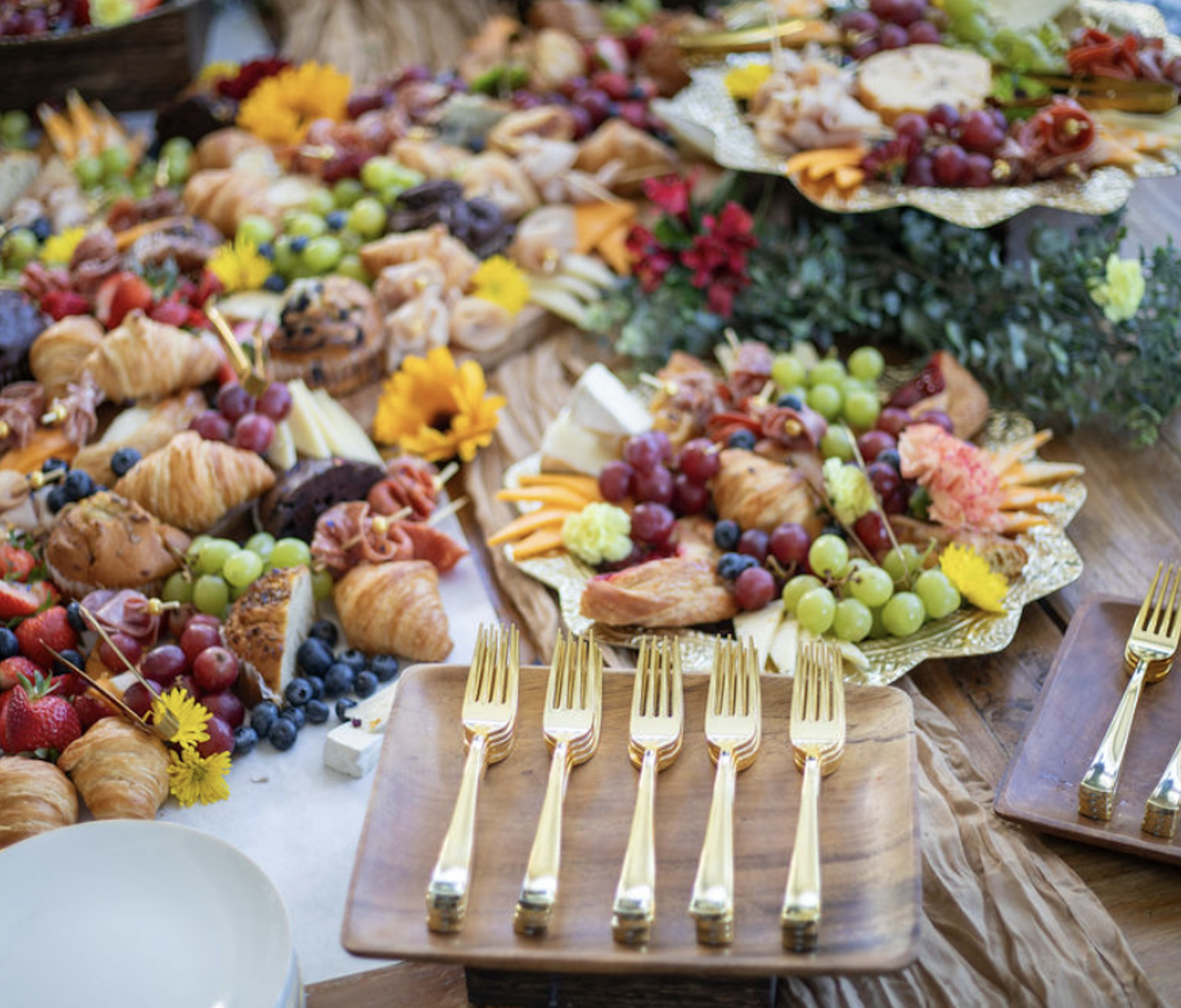 A table set for a celebration with a variety of finger foods and snacks, including grapes, strawberries, cheese, and pastries, arranged on decorative platters, with gold forks and a wooden serving tray.