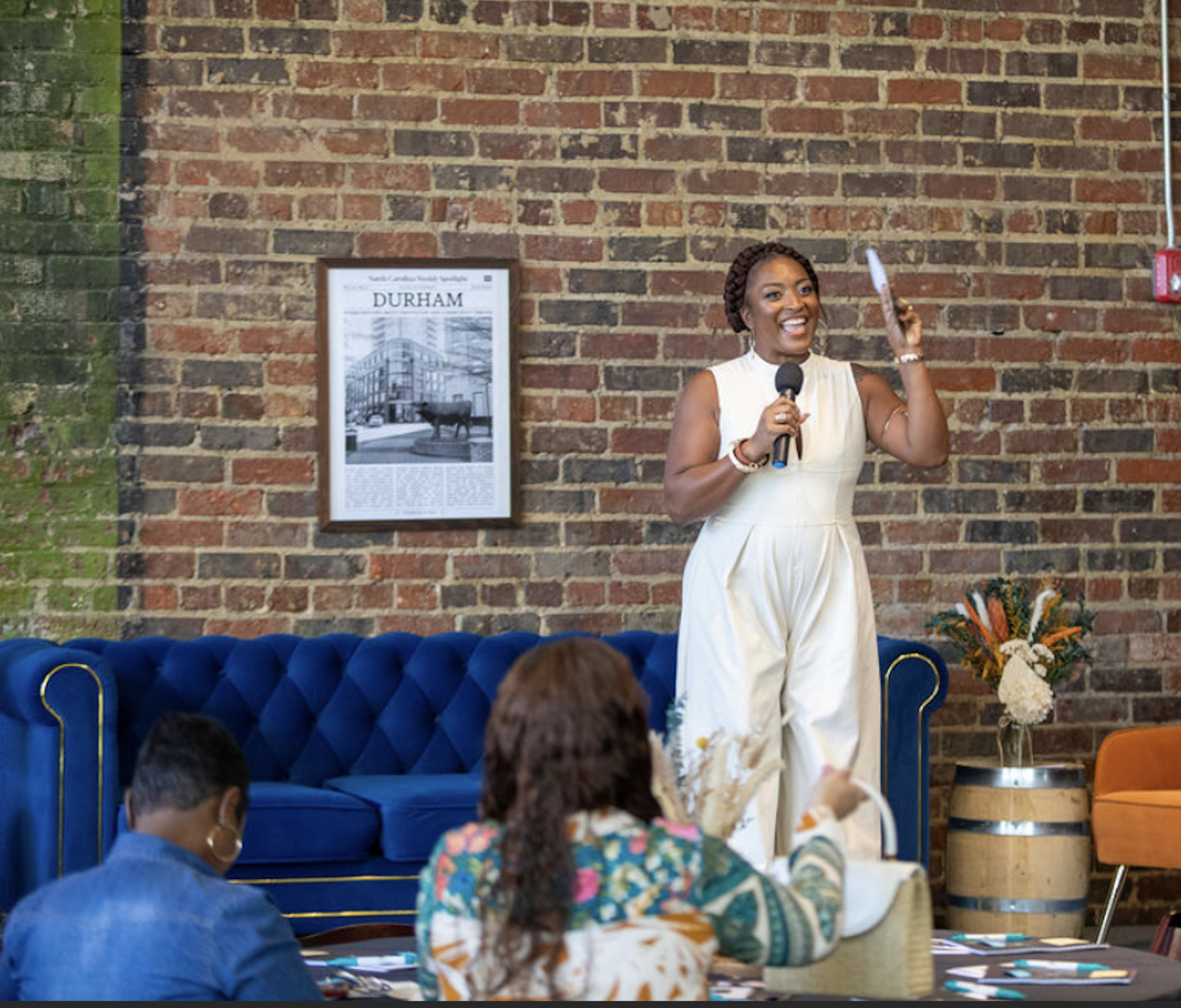 A woman standing and smiling while speaking into a microphone during a presentation or speech in a cozy, modern venue with a brick wall background, framed article about Durham, and floral decor.