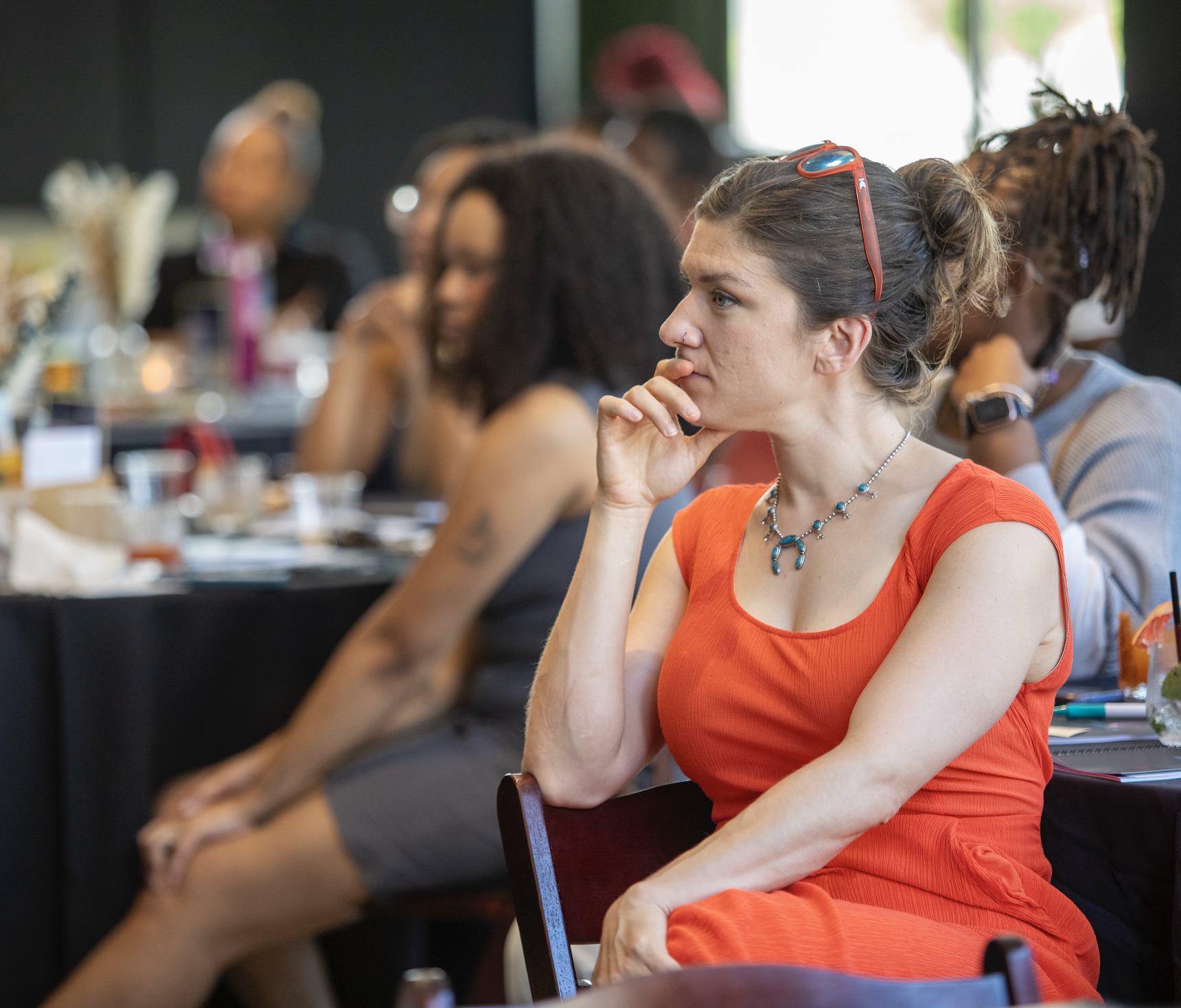 A woman in an orange dress with red sunglasses on her head and a blue necklace sitting at a table during a meeting, with other women in the background.