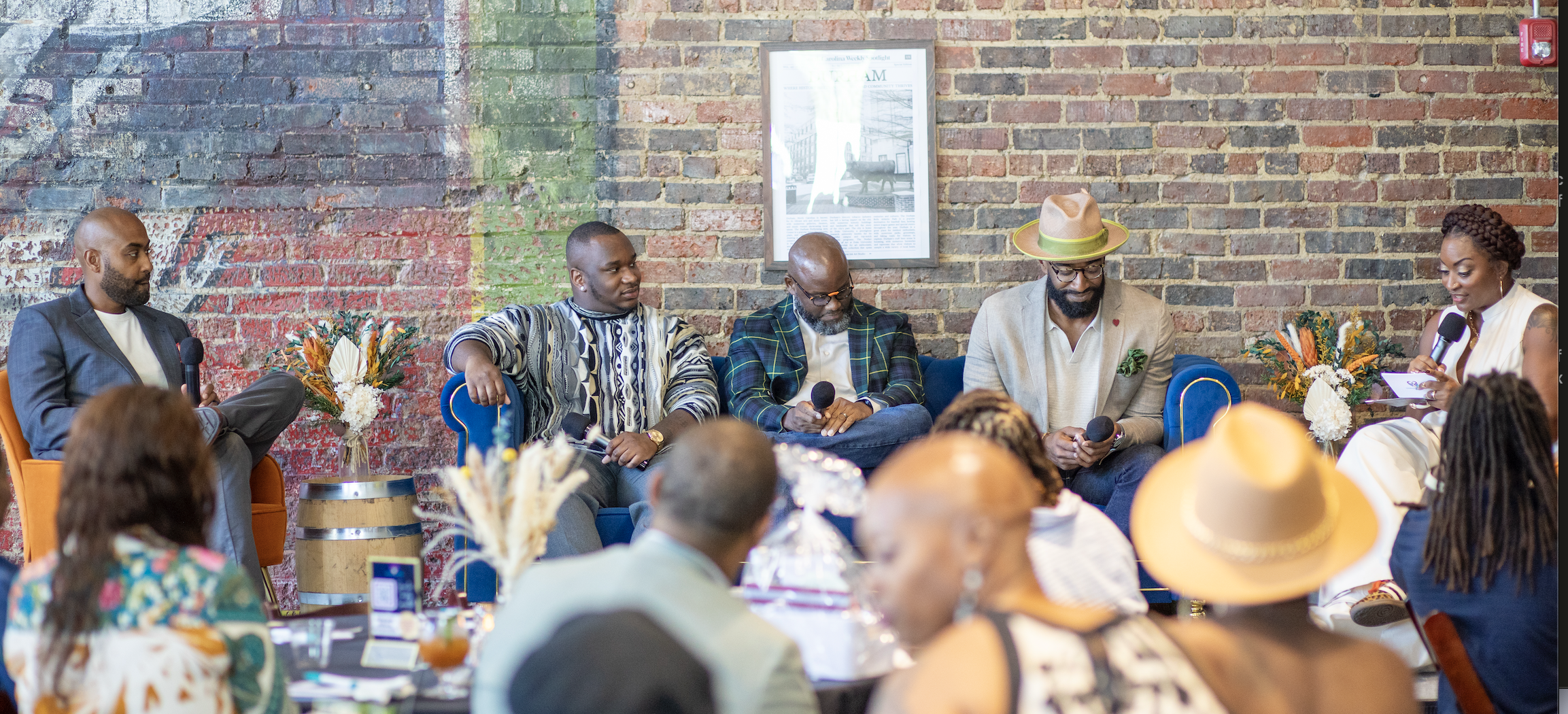 A panel of five people participating in a discussion or interview at an event, sitting on a sofa with a brick wall background, with an audience in front. The panel includes four men and one woman, each holding a microphone, and there are floral arran