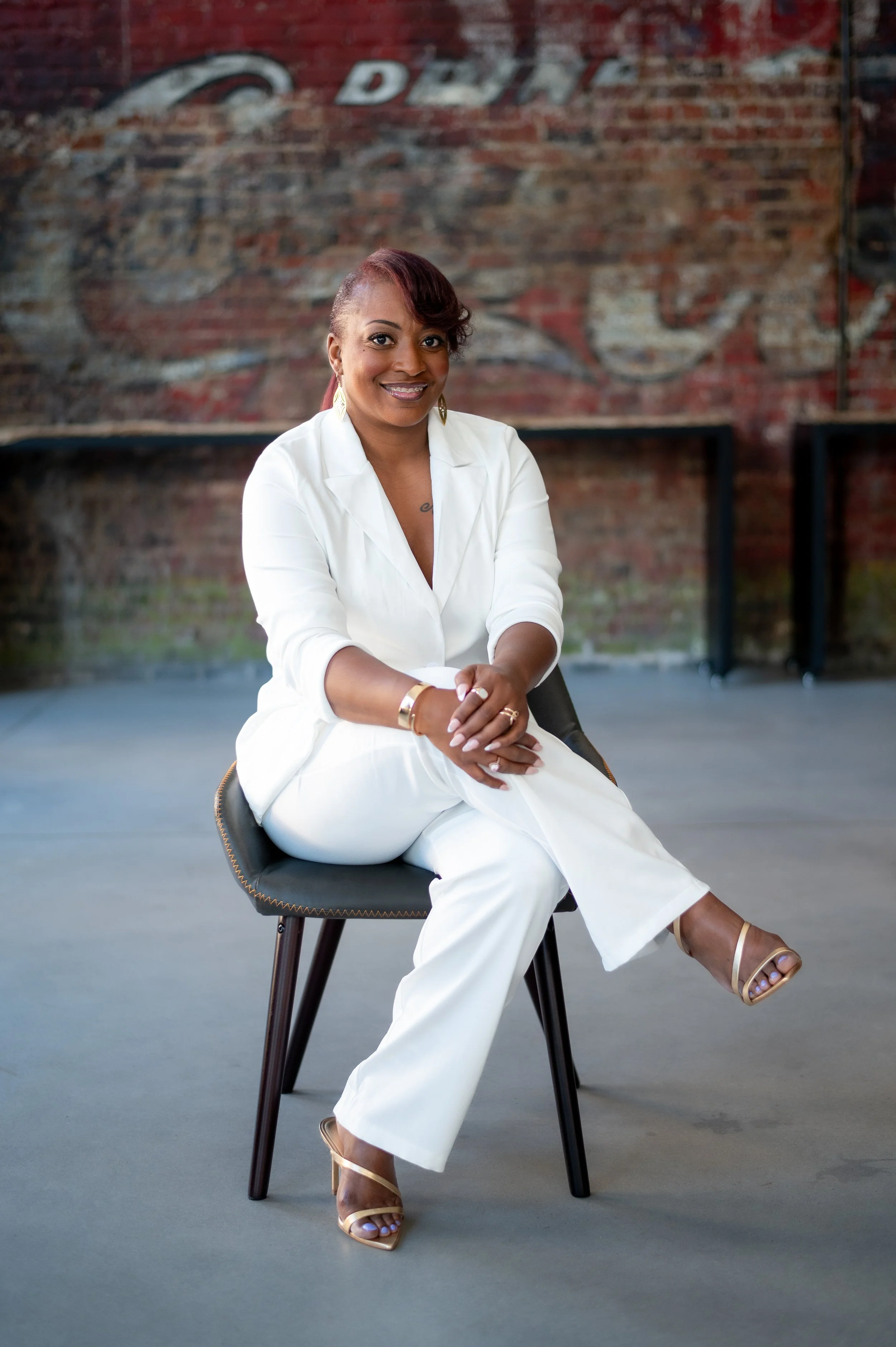 A woman with short, dark red hair styled to one side, sitting on a modern black chair, smiling, wearing a white suit with gold heels, with a background featuring exposed brick wall and a vintage sign.