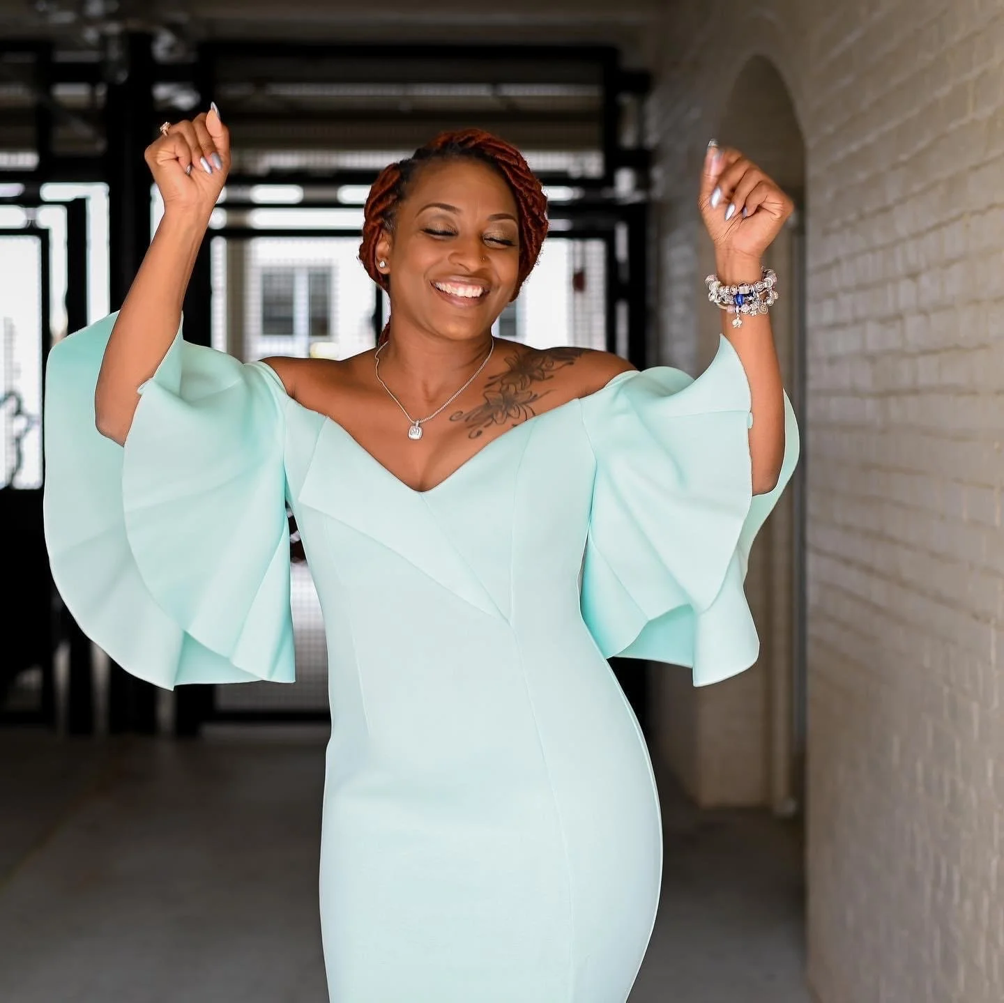 A woman with short curly hair in a mint green dress with ruffled sleeves, smiling and raising her arms in a celebratory pose indoors.