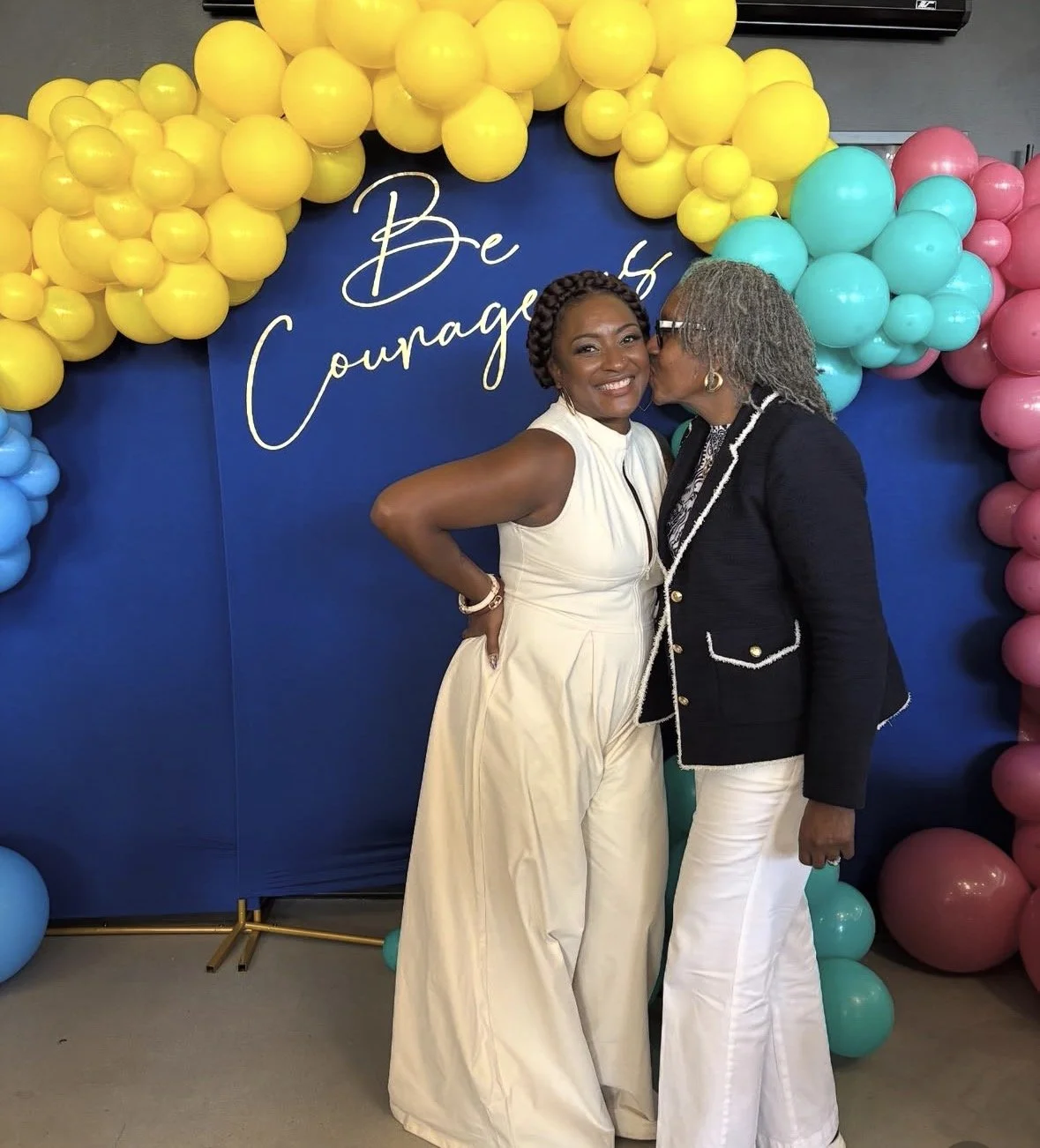 Two women smiling and sharing a kiss on the cheek in front of a colorful balloon arch and a blue backdrop with the words "Be Courageous" written on it.
