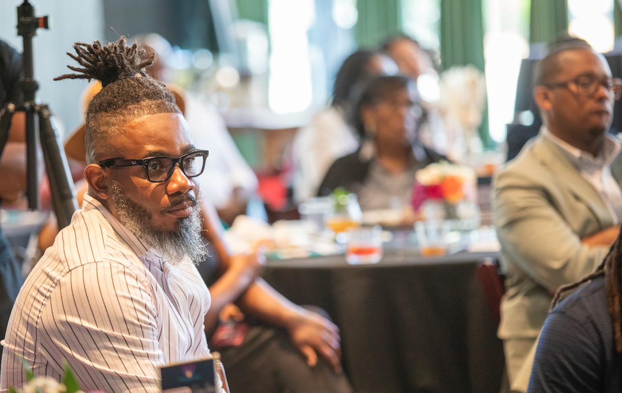 A man with glasses, a beard, and dreadlocks tied in a bun, wearing a striped shirt, sitting attentively at a conference or meeting.