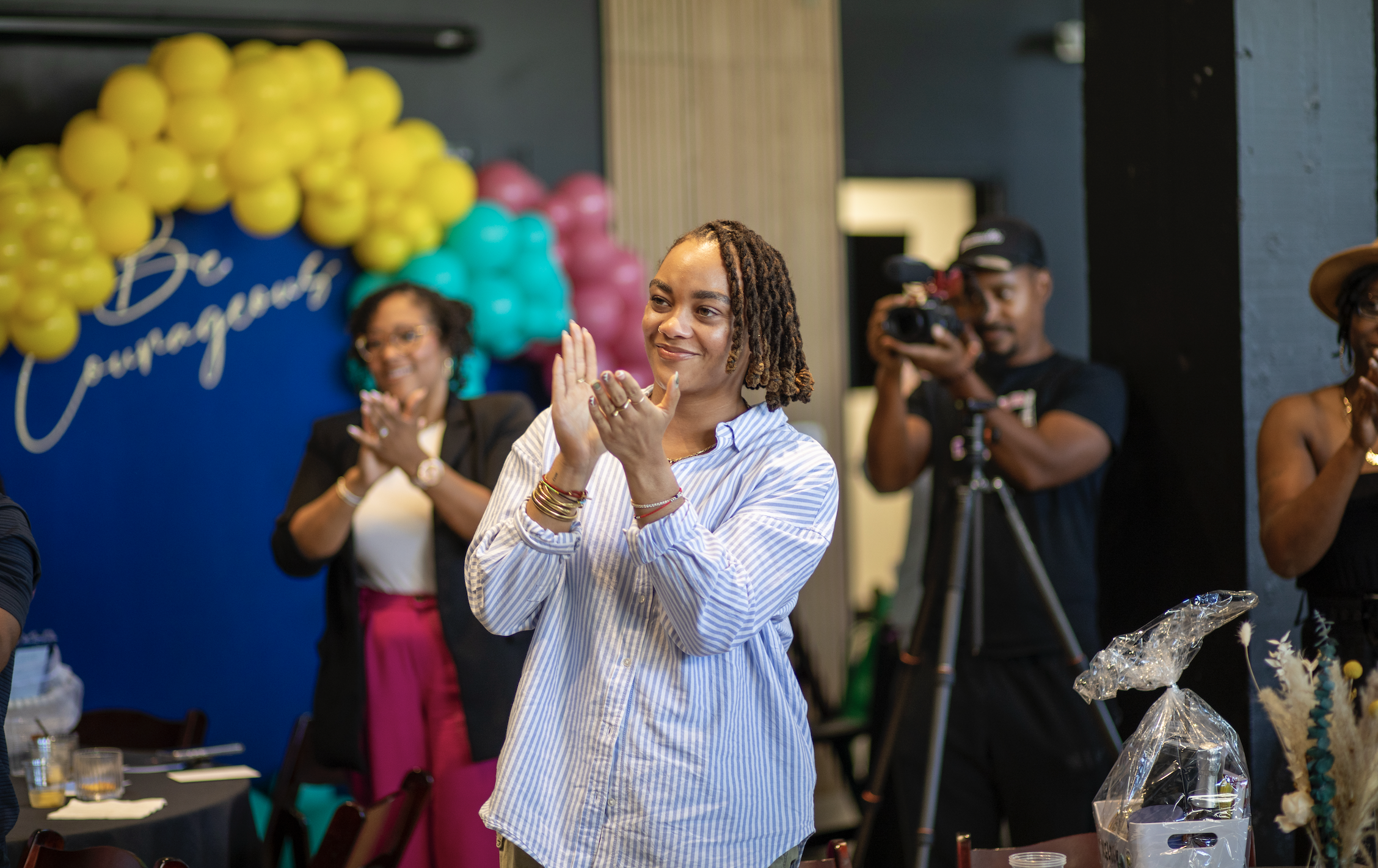 Women clapping at a celebration event with colorful balloons and a 'Congratulations' sign in the background.