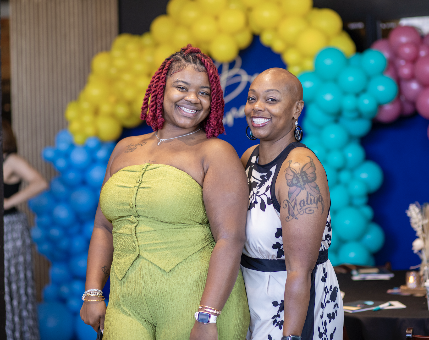 Two women smiling at a celebration, standing in front of colorful balloons with yellow, blue, turquoise, and pink colors, wearing dresses and jewelry.