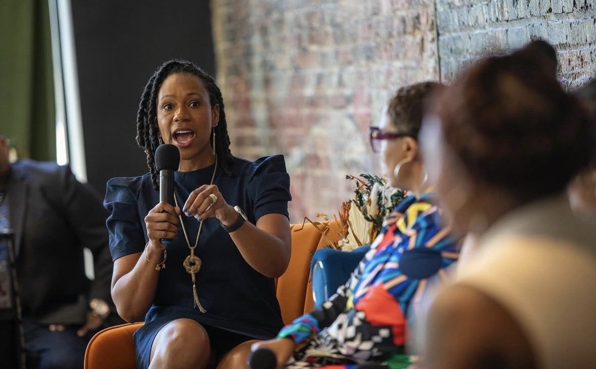 A woman with dreadlocks speaking into a microphone during a panel discussion, seated on an orange chair, with women listening to her, set against a brick wall.