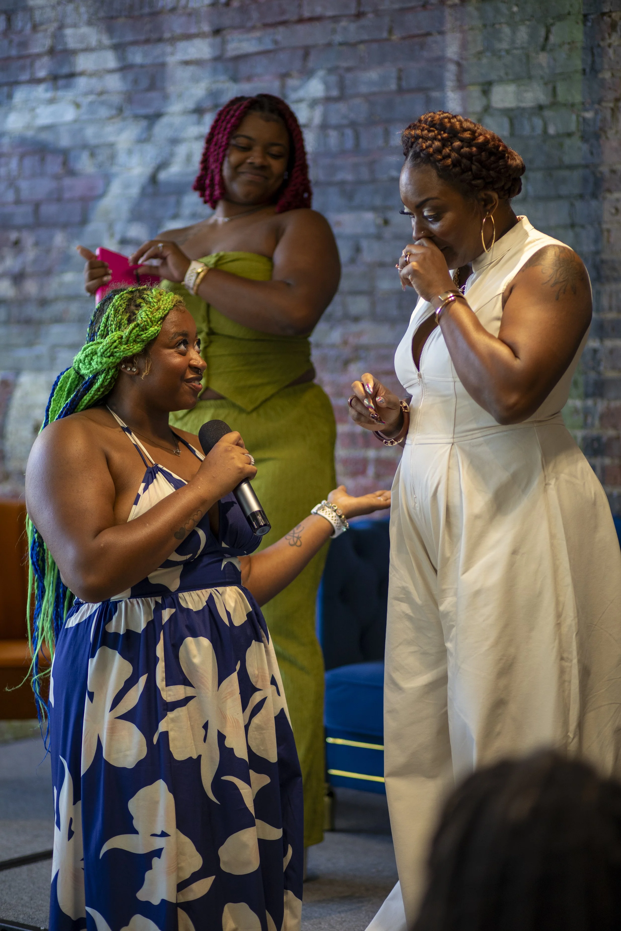 Three women engaged in a heartfelt moment, with one woman holding a microphone and gesturing towards a woman in a beige jumpsuit, while a third woman in a green dress stands in the background, smiling and holding a pink item.