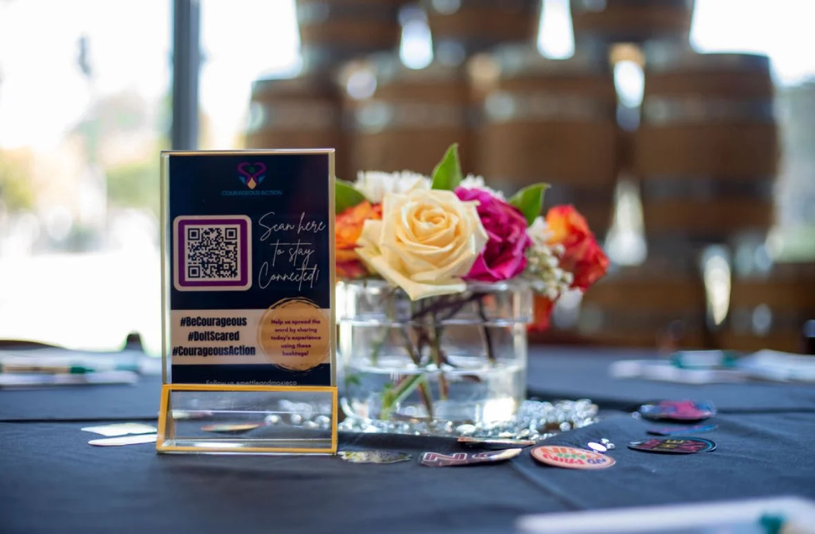 A table with a flower arrangement in a glass vase, a sign with a QR code and motivational message, and scattered buttons or small decorations, in a well-lit indoor setting.