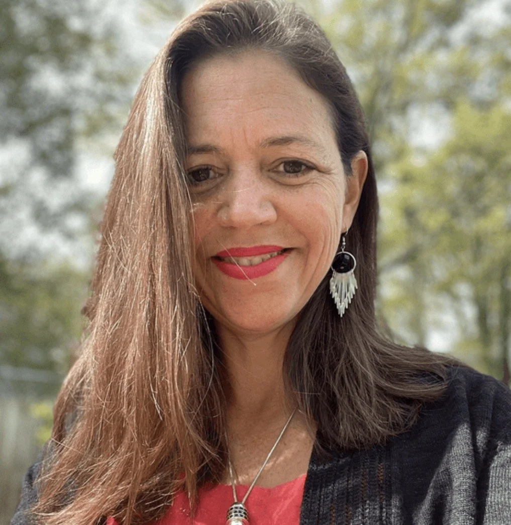 A woman with long brown hair, wearing red lipstick, earrings with black and silver accents, and a necklace, outdoors with blurred trees in the background.