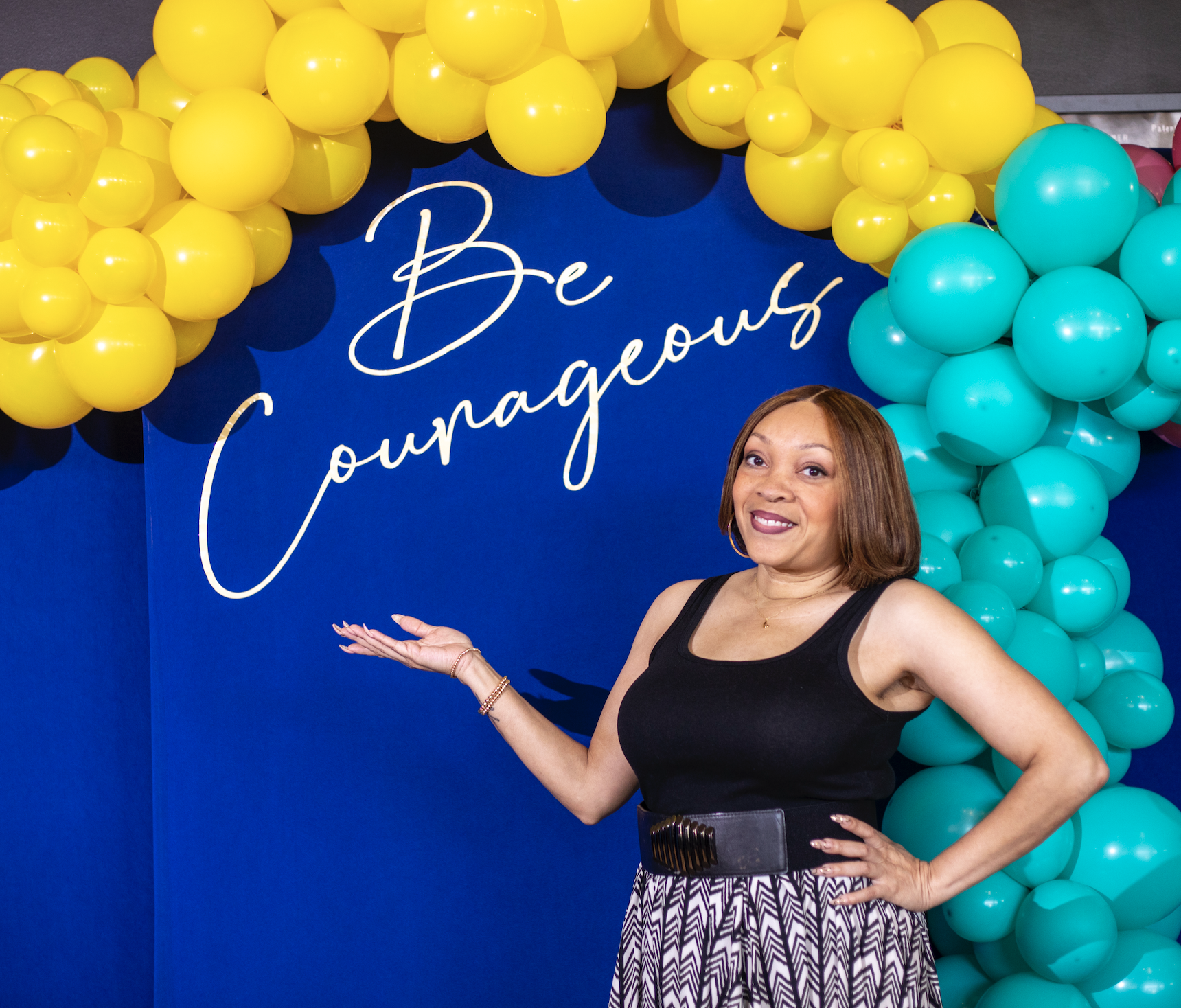 A woman standing in front of a blue backdrop decorated with yellow and teal balloons, with the words 'Be Courageous' written on the backdrop. She is wearing a black sleeveless top and patterned pants, smiling with one hand on her hip and the other ge