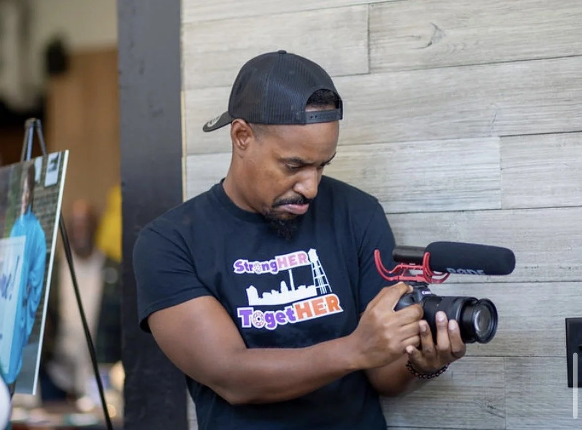 A man wearing a black cap and t-shirt with the words "StrongHER TargetHER" is holding a camera with a mounted microphone, standing against a light wooden wall.