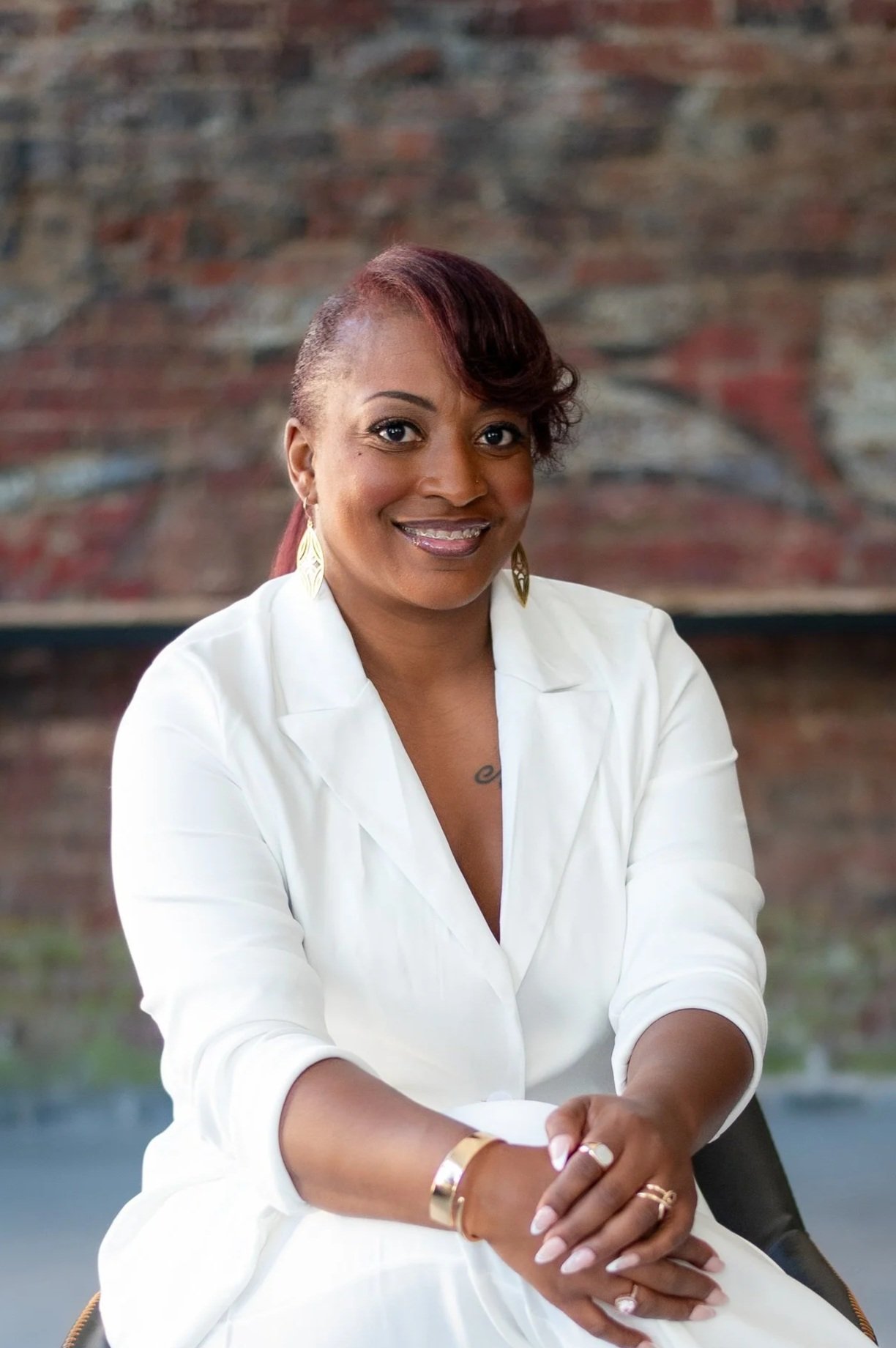 A smiling woman with braided hair, wearing a white outfit and gold jewelry, sitting in front of a brick wall.