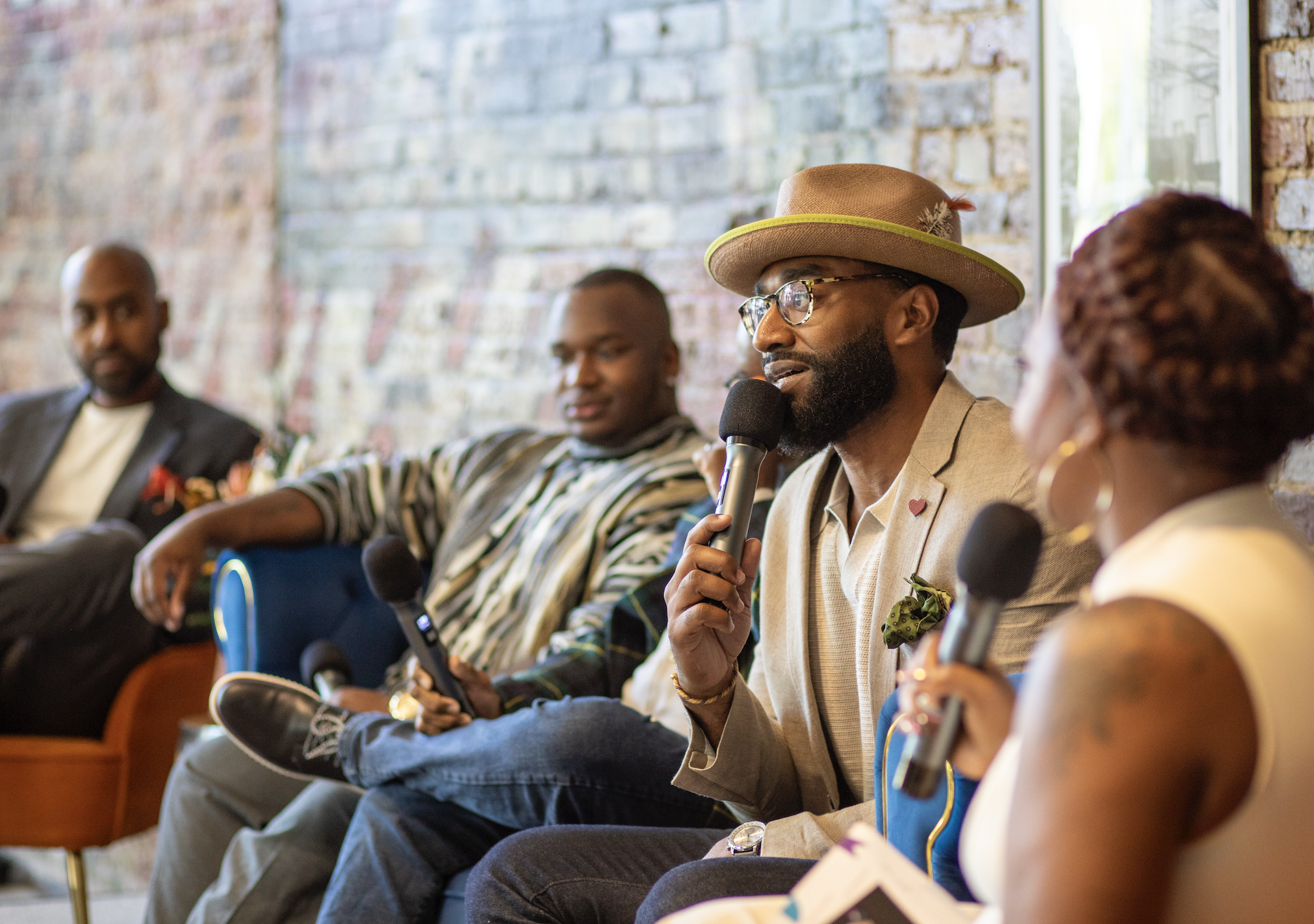 A diverse group of four people participating in a panel discussion, with one person speaking into a microphone, sitting against an exposed brick wall.