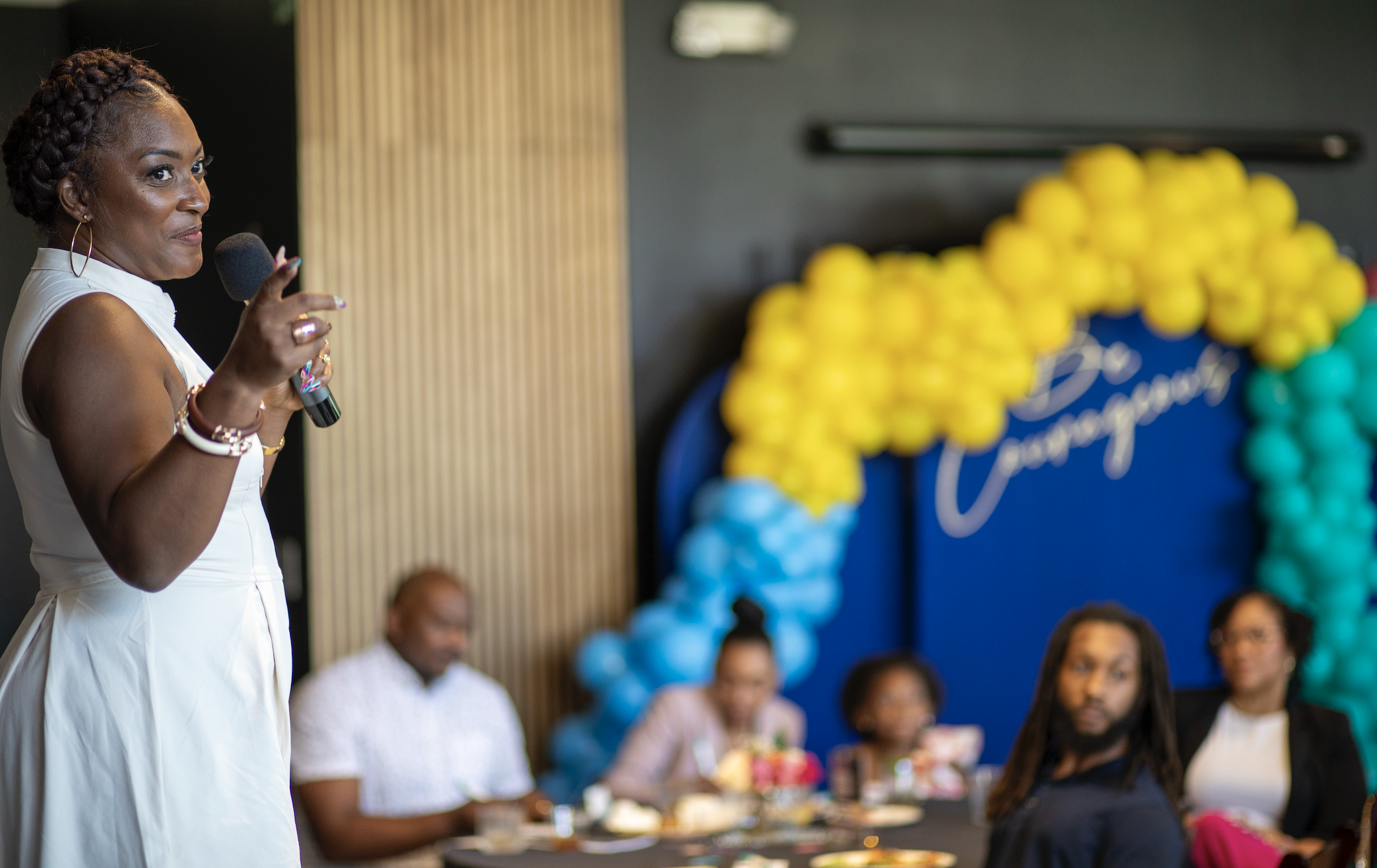 A woman in a white dress holding a microphone at a celebration event with a balloon arch in the background.