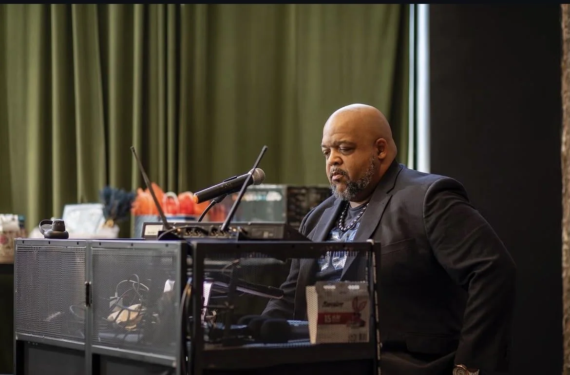 A man with a beard sits at a piano, with a microphone in front of him, in a room with green curtains.