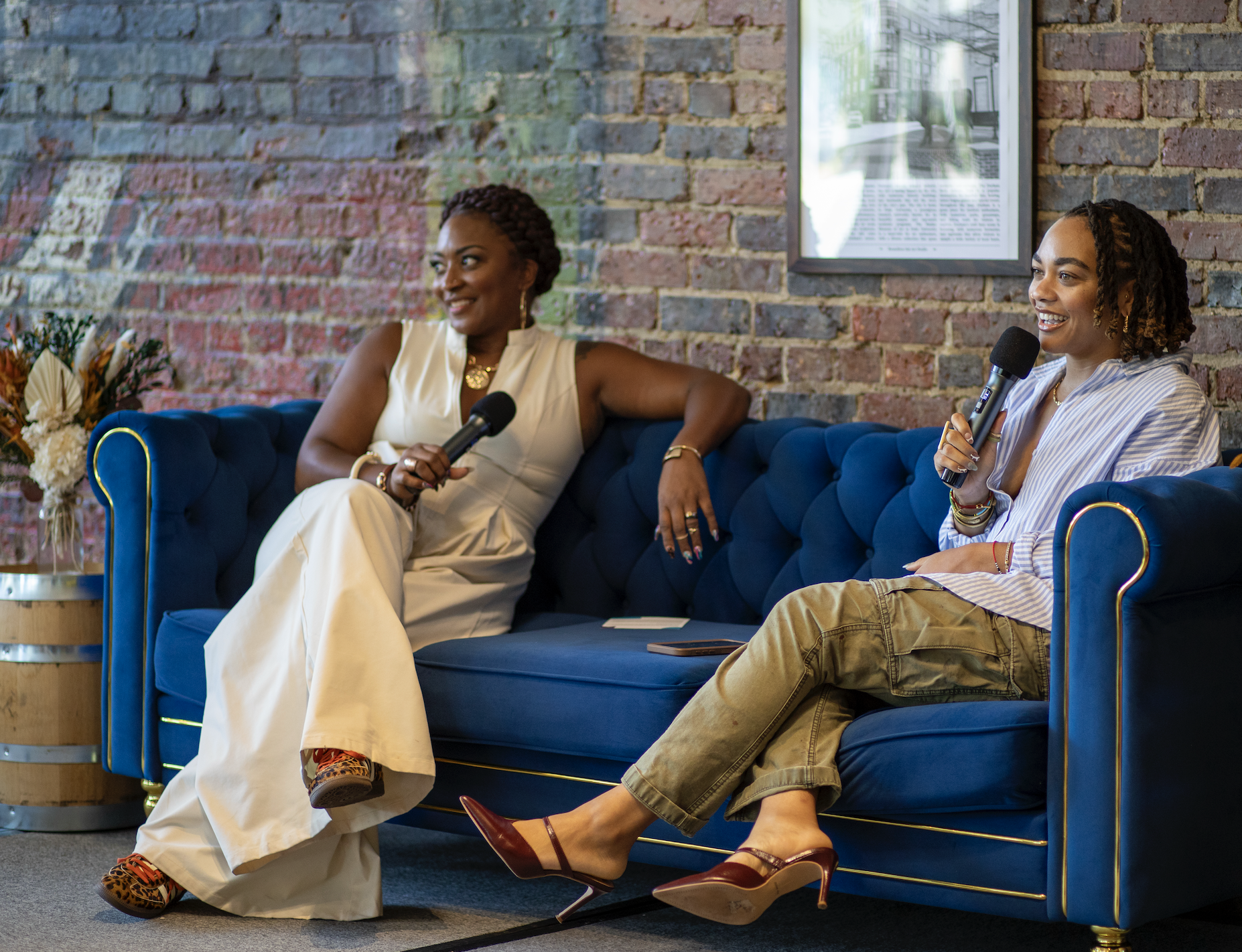 Two women sitting on a blue velvet sofa, each holding a microphone, engaged in a conversation. The woman on the left is wearing a sleeveless beige outfit, and the woman on the right is dressed in a light striped shirt and cargo pants. They are in a r