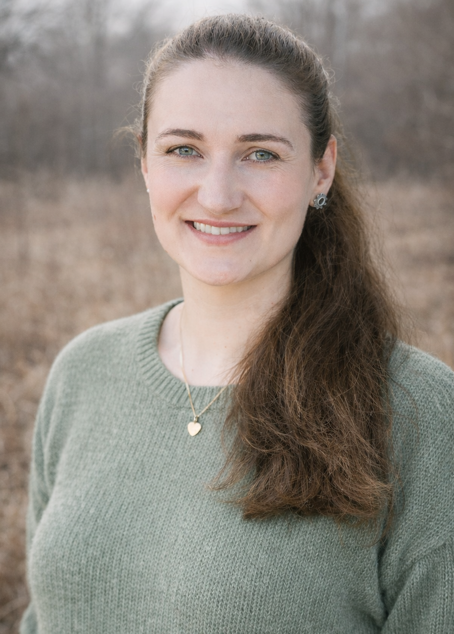 Lächelnde junge Frau mit braunen, gewellten Haaren, blauen Augen, grüner Kleidung und Schmuck, im Freien mit unscharfem Hintergrund.