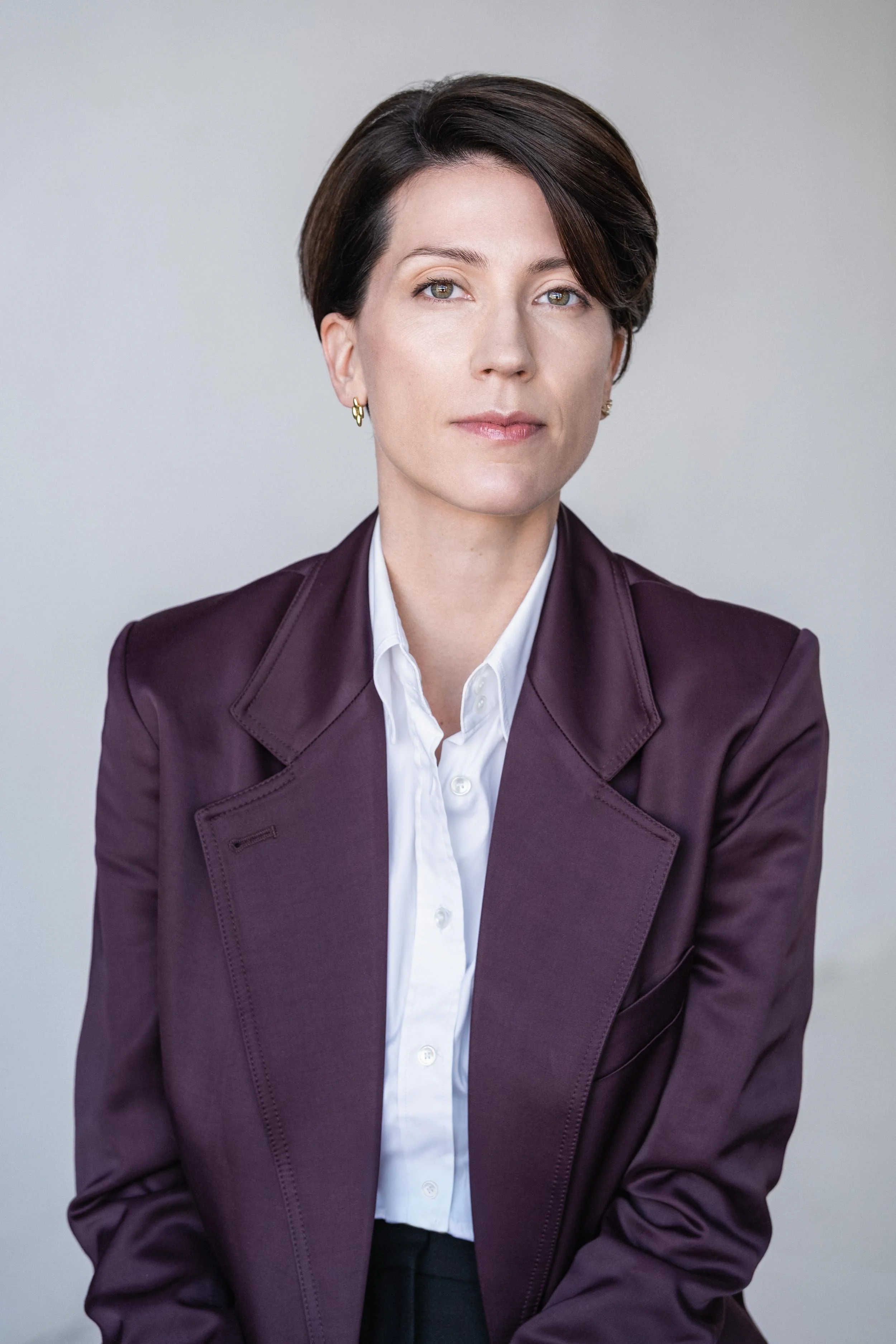 Portrait d'une femme portant une veste violette et une chemise blanche, avec des cheveux courts bruns et un fond gris clair.