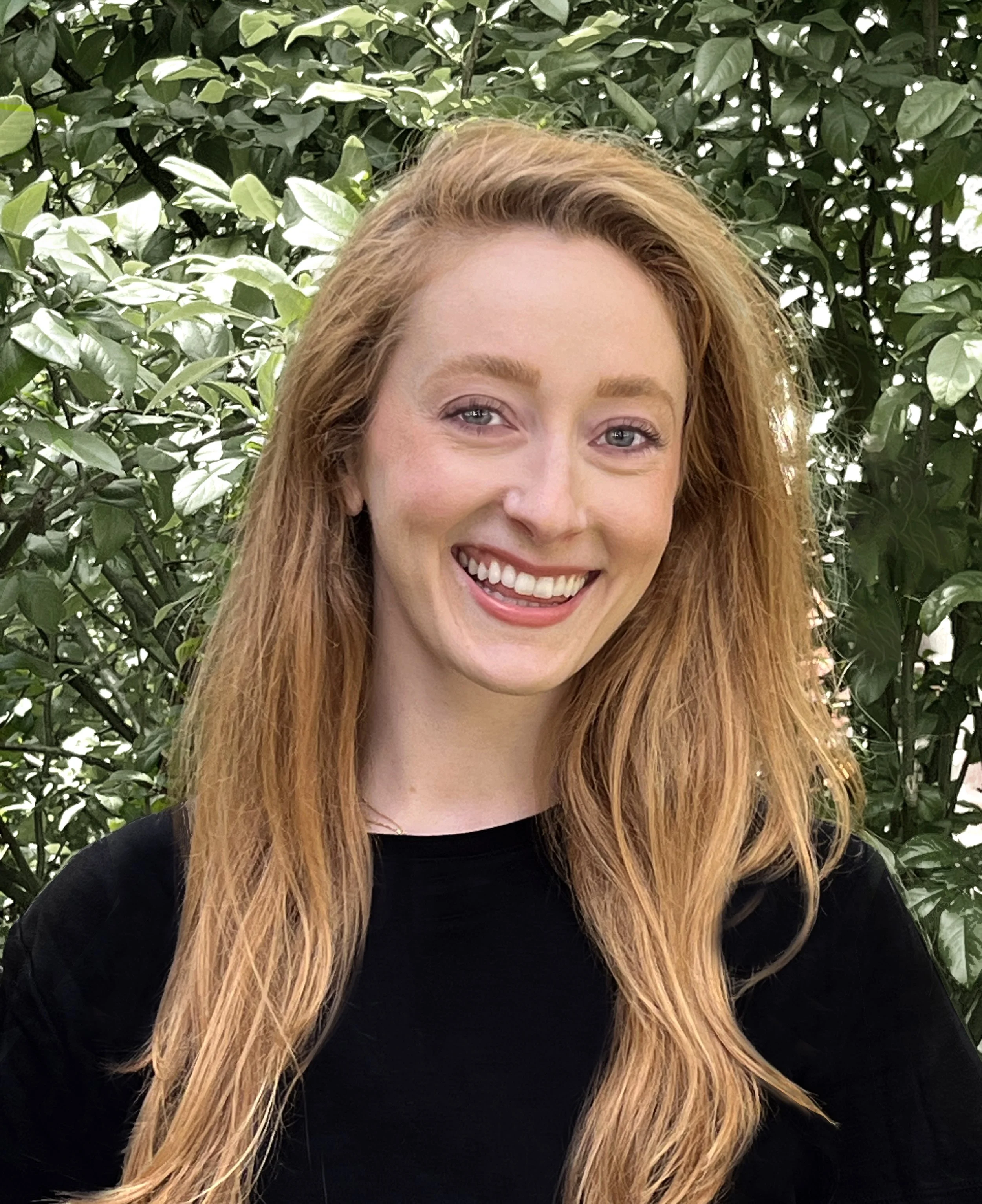 A smiling woman with long red hair and blue eyes standing outdoors in front of green leafy foliage.