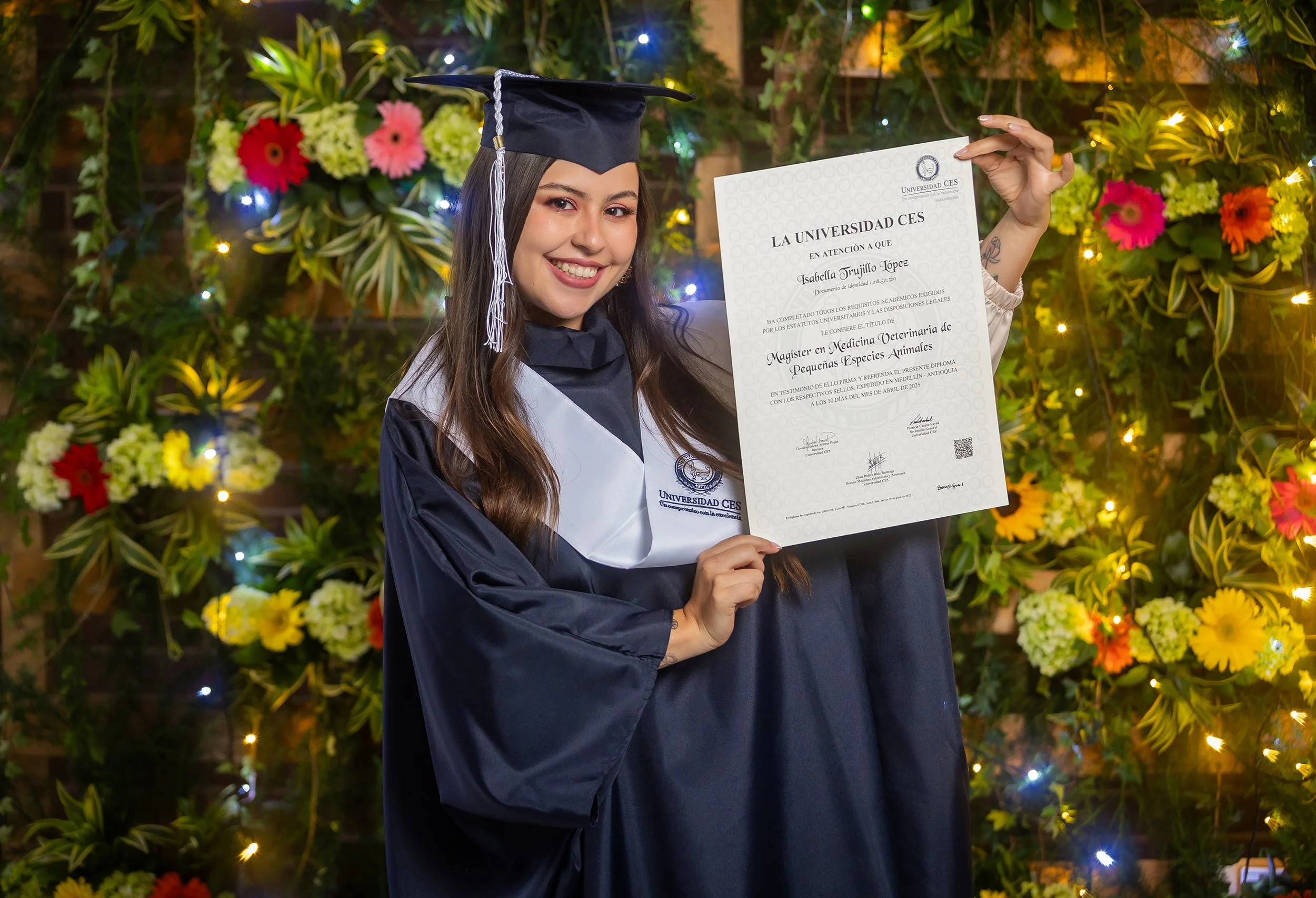 Joven graduada con toga y birrete, sosteniendo un diploma en un fondo decorado con flores y luces.