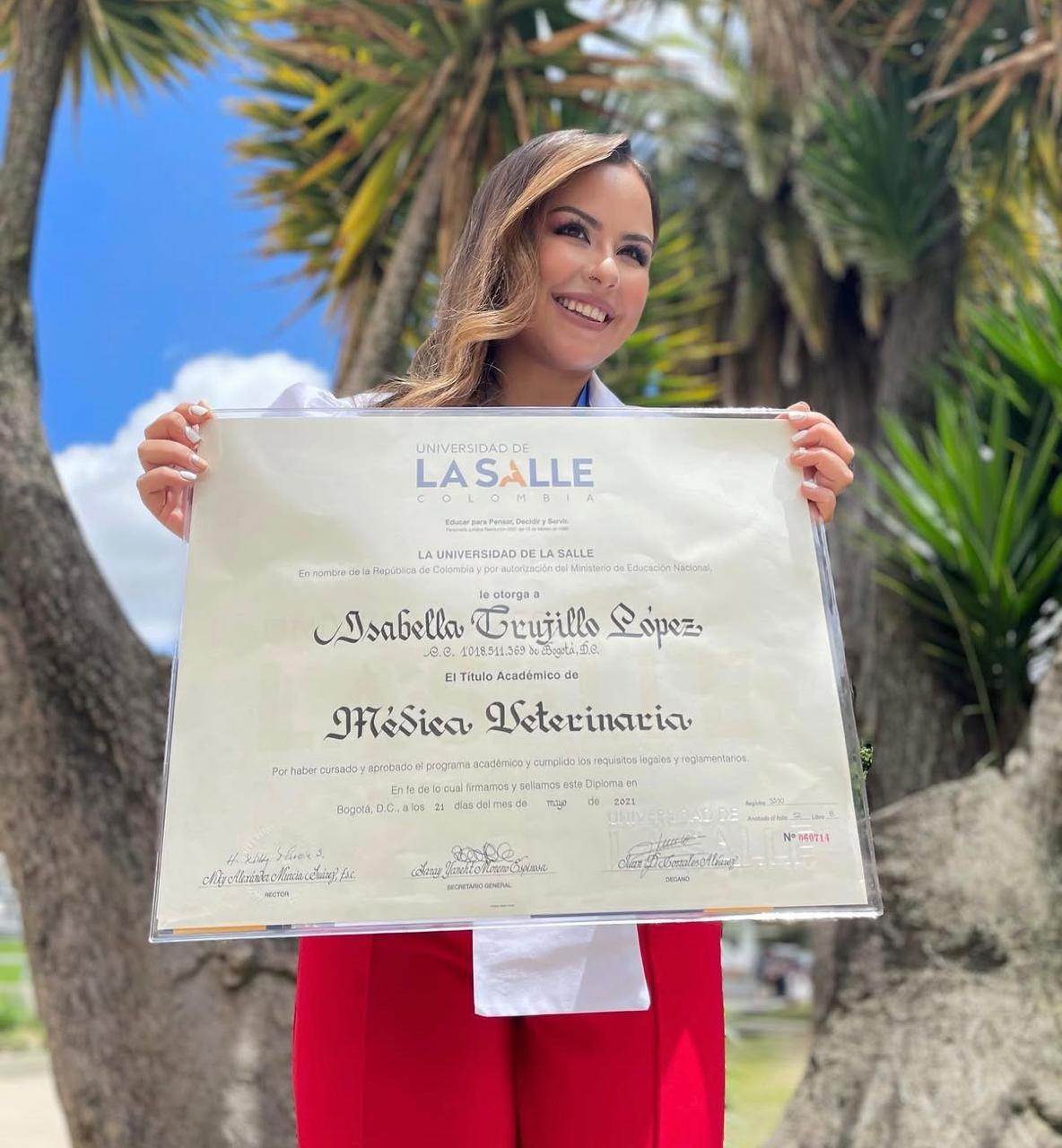 Joven mujer en ceremonia al aire libre sosteniendo título universitario en ciencias veterinarias. Fondo con árboles y cielo despejado.
