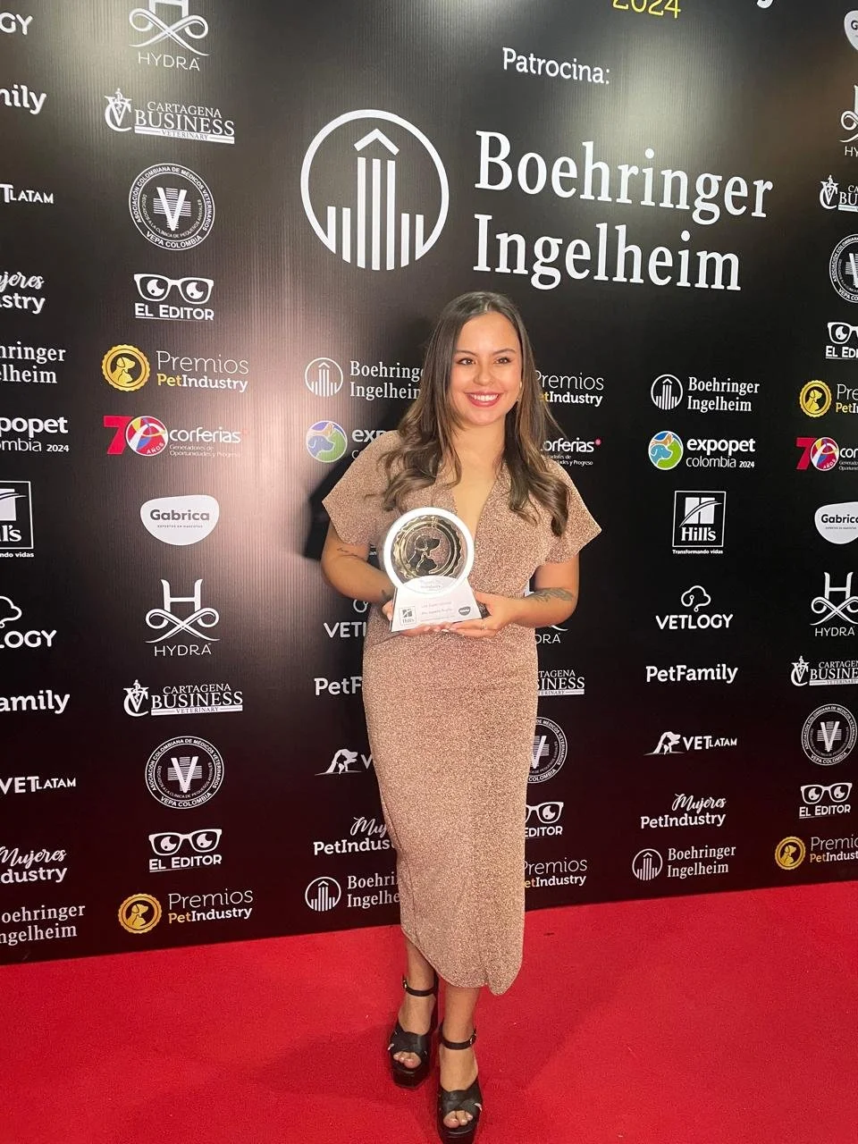 Mujer sonriendo y sosteniendo un trofeo en un evento de premiación con fondo negro y logotipos.