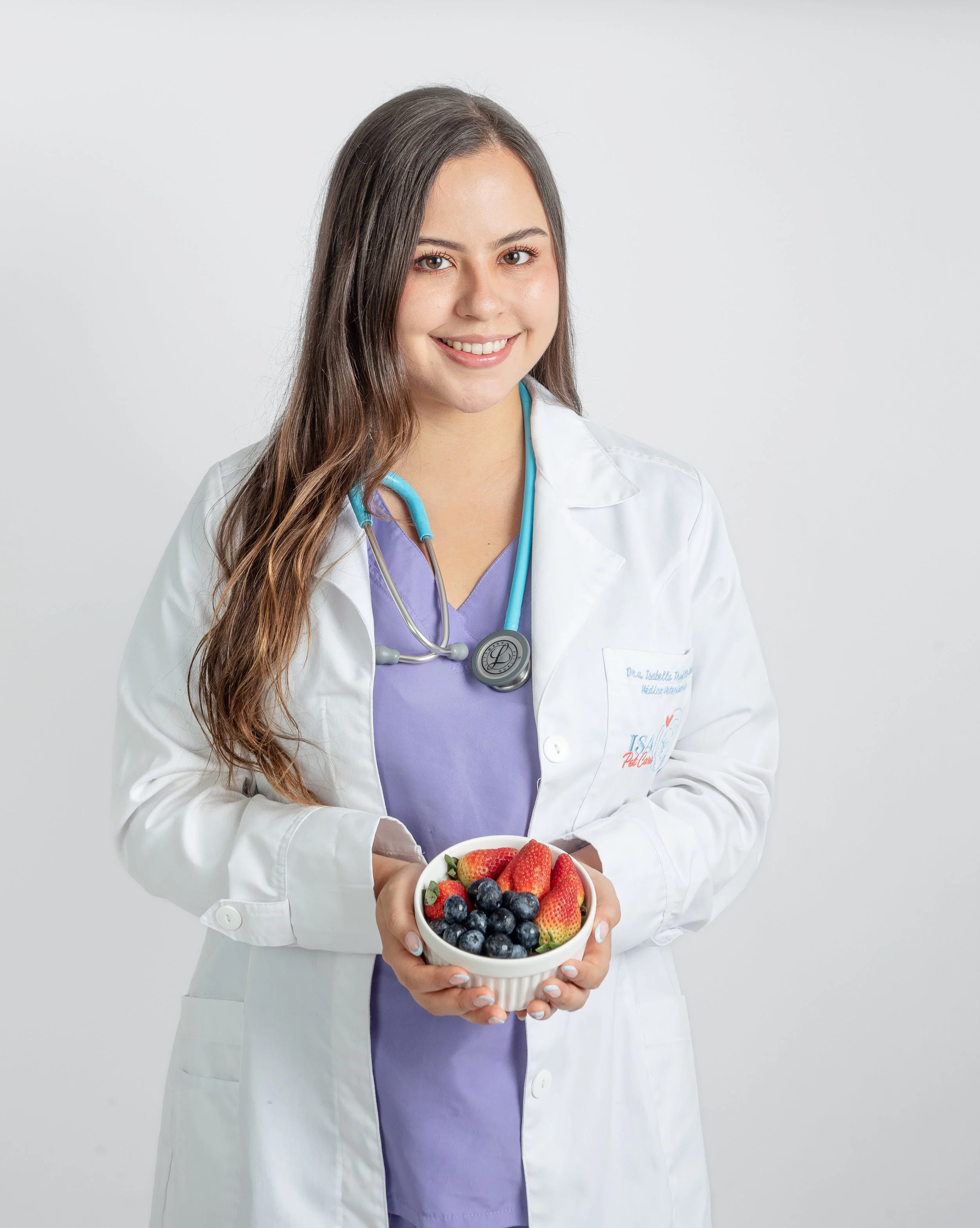 Médica sonriendo, usando bata blanca y estetoscopio, sosteniendo un tazón con fresas, arándanos y frambuesas.