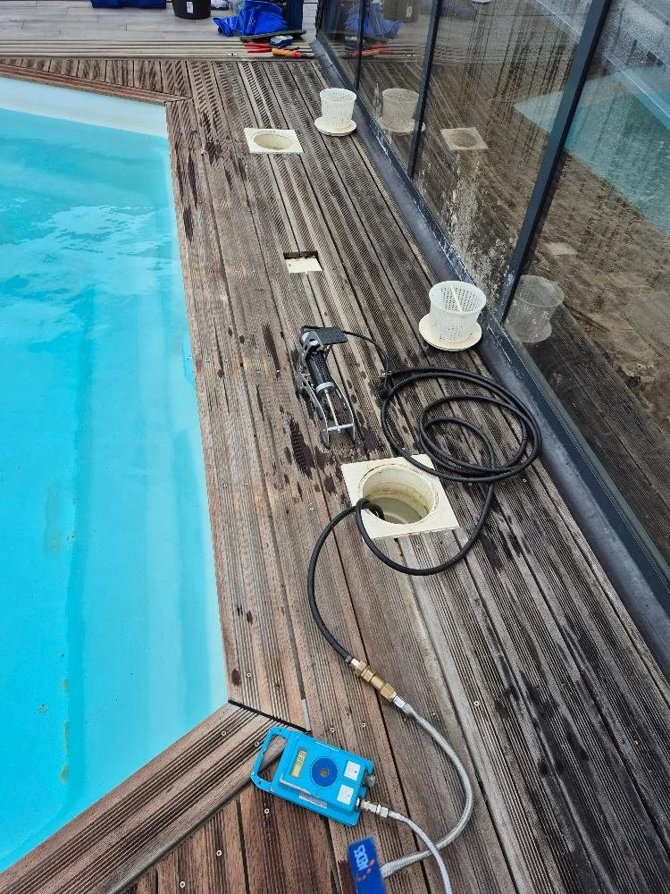 Outils de nettoyage pour piscine sur un patio en bois, dont un pistolet à haute pression, un xylème d'entretien, et des enrouleurs de câble, près d'une piscine.