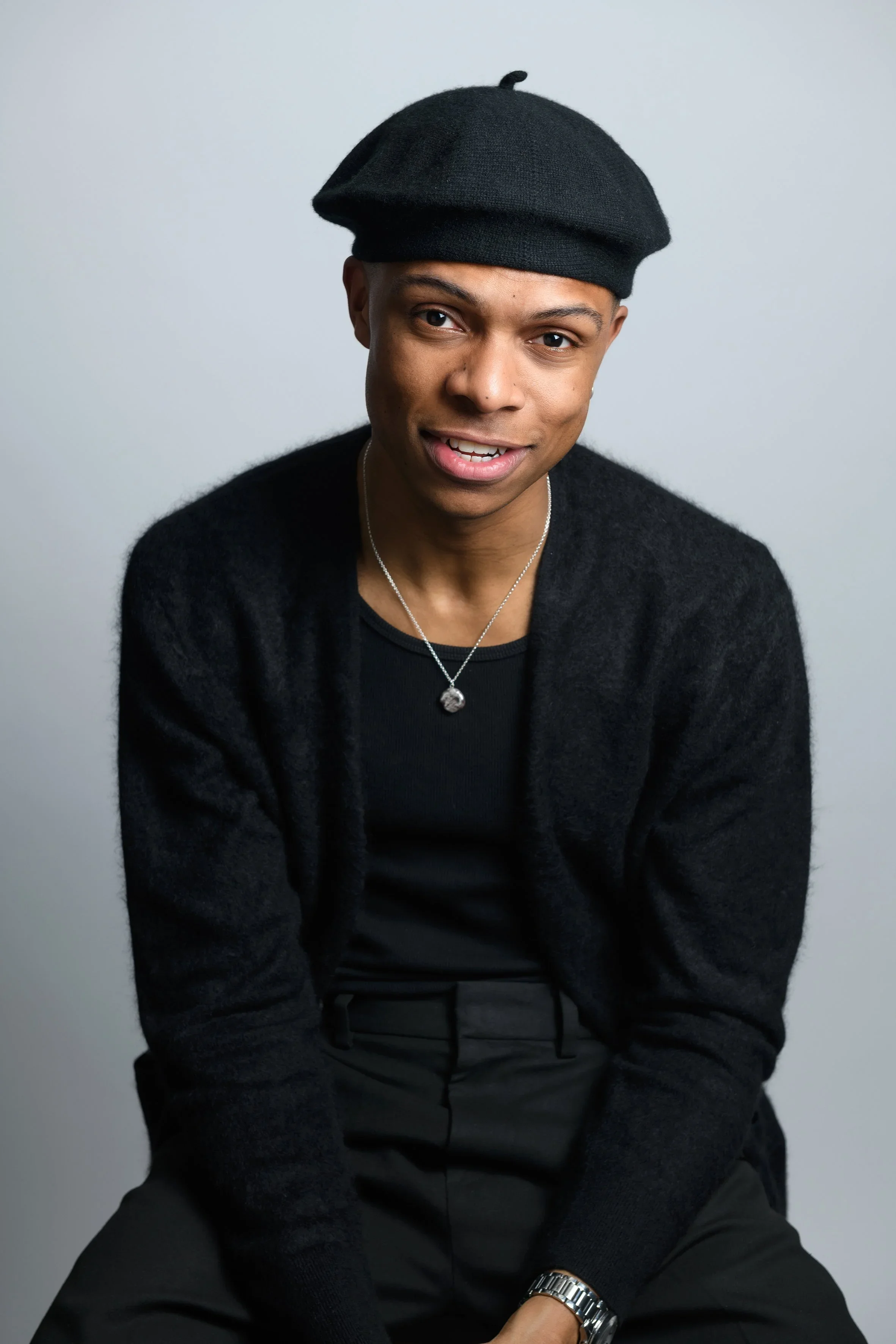 Comedian Fab Goualin wearing a black beret, black sweater, and black pants sitting against a plain white background.