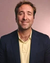 Comedian Joe Bor with light brown hair, a slight beard, and a warm smile, wearing a dark blazer over a light yellow shirt, standing against a plain background.