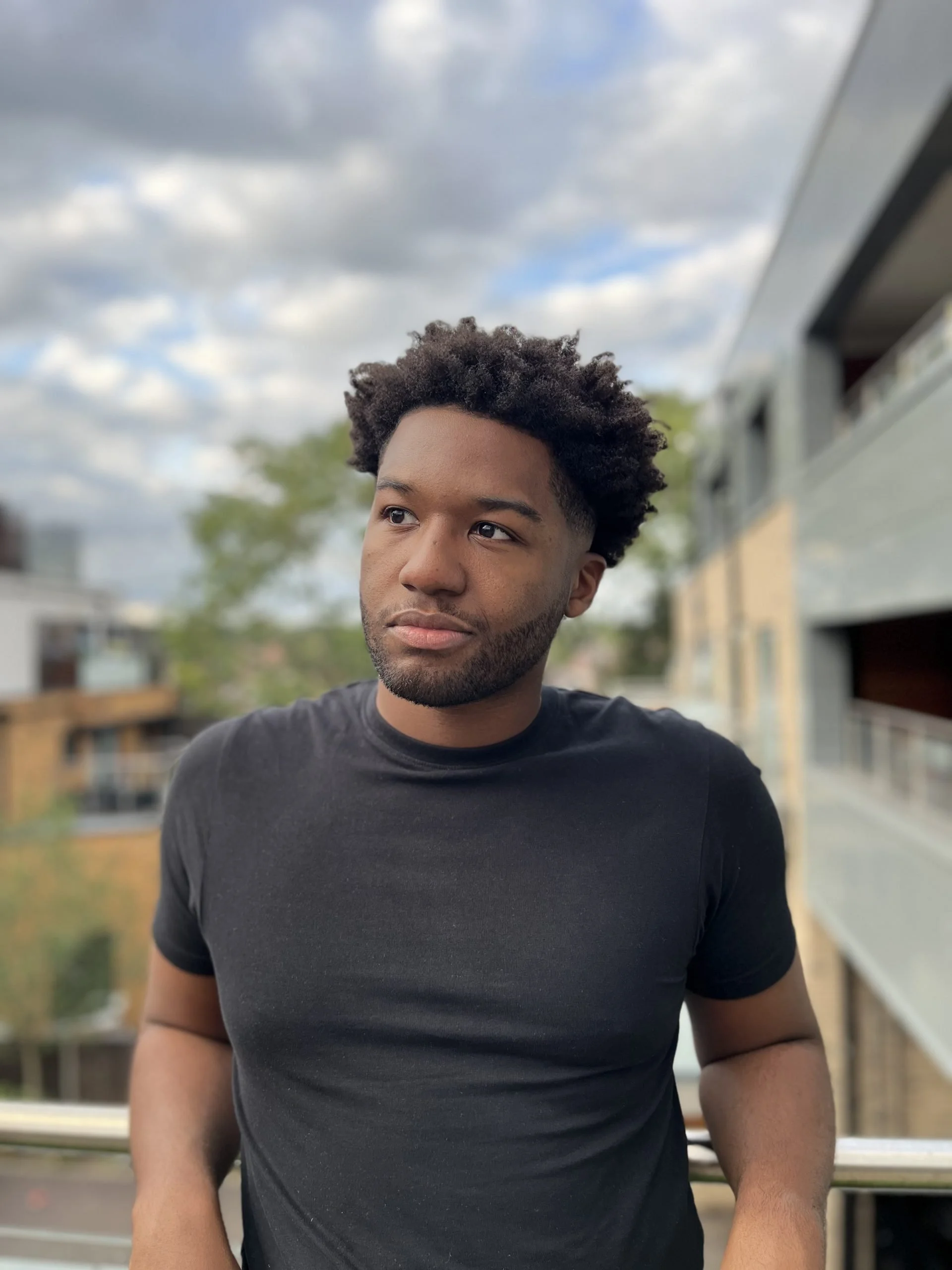 Comedian Wes Defoe with dark curly hair and a beard, wearing a black t-shirt, standing outdoors on a balcony with cloudy sky and buildings in the background.