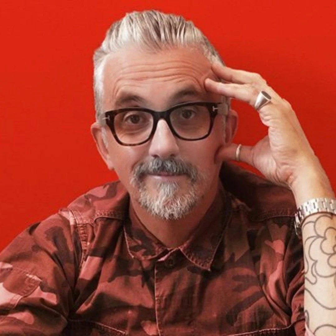 Comedian Markus Birdman with gray hair, glasses, and a mustache, wearing a red patterned shirt, sitting against a red background, posing with his hand on his forehead and looking into the camera.