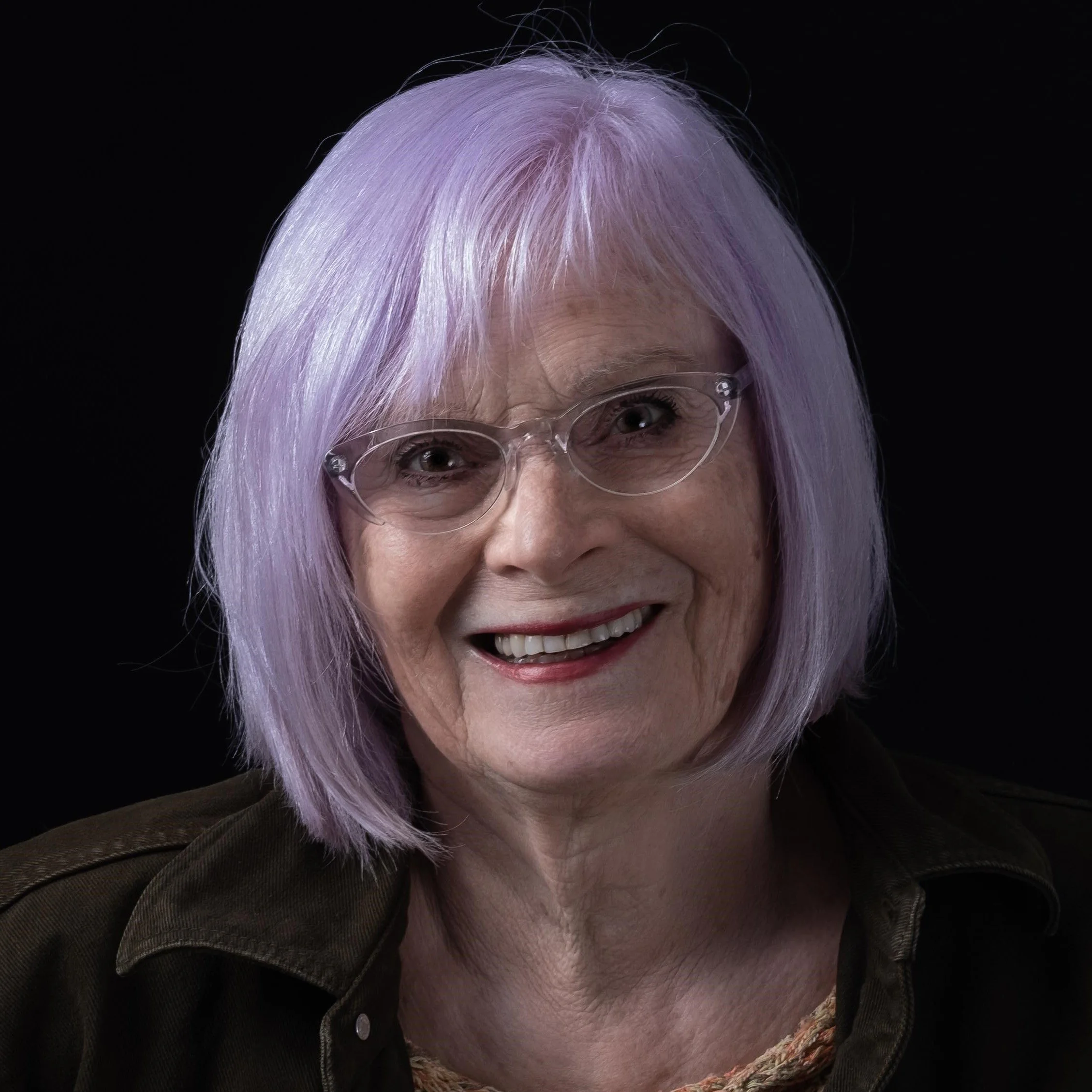 Portrait of a woman, Hils Jago, with light purple hair, glasses, and a black background.