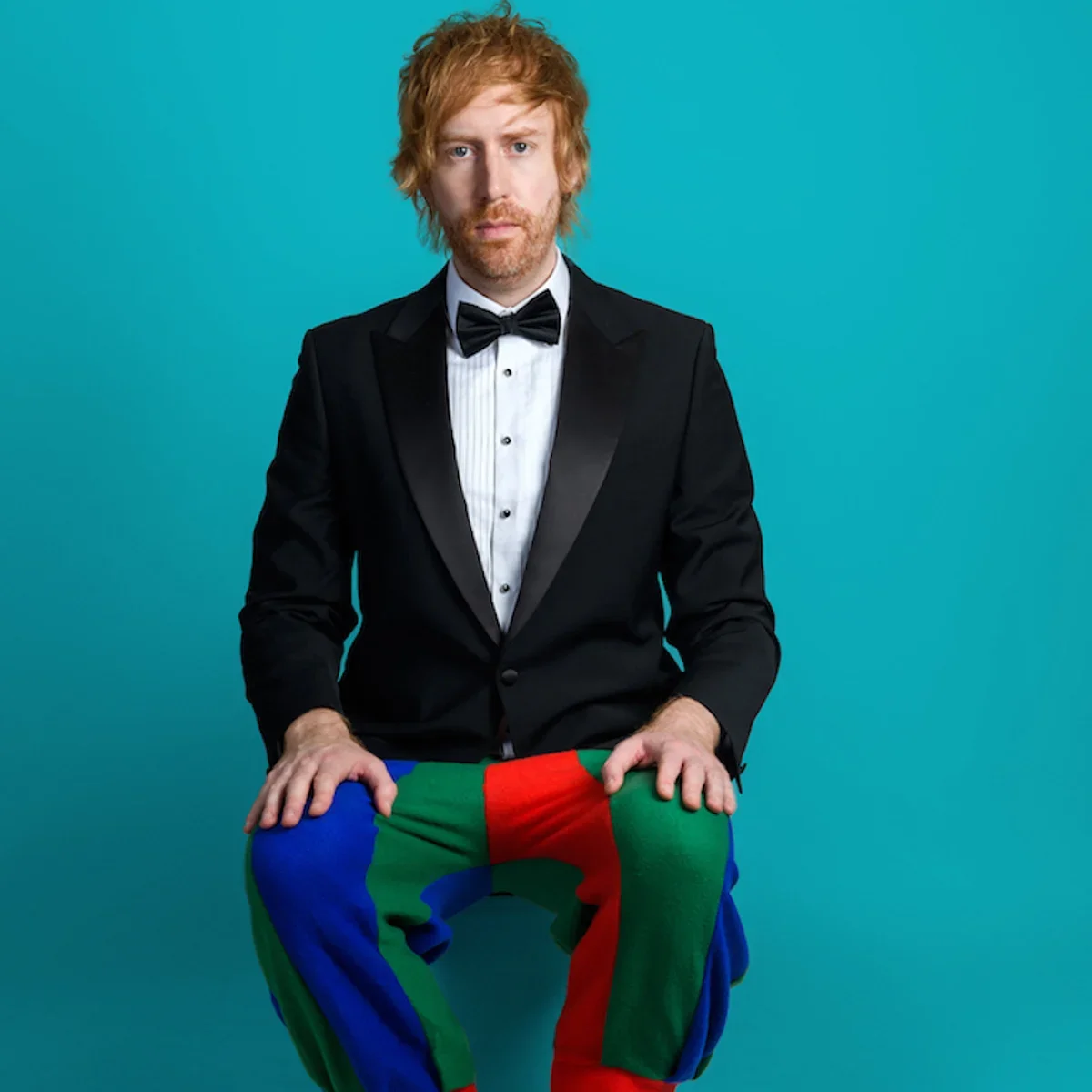 A man with ginger hair and beard dressed in a tuxedo with a bow tie, sitting against a blue background, holding colorful patchwork pants.