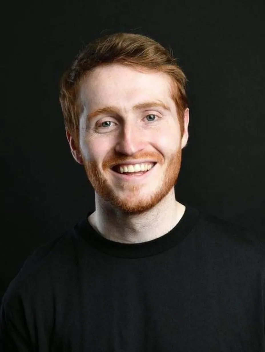 Comedian Ali Woods with red hair and beard, smiling, wearing a black shirt against a black background.